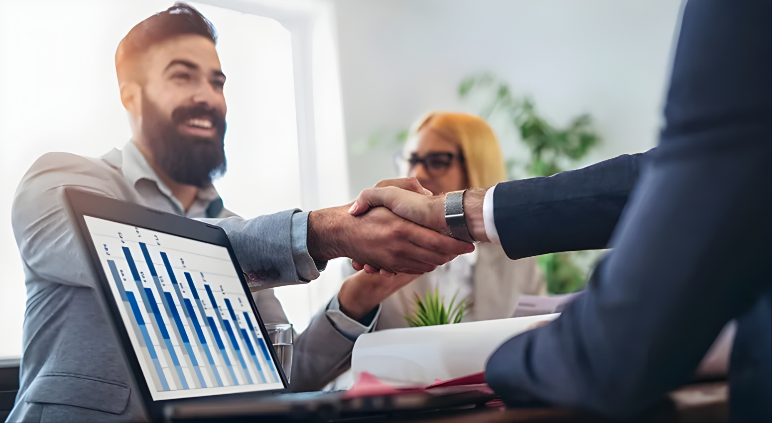 Two business professionals shaking hands during a meeting with financial charts visible on a laptop.
