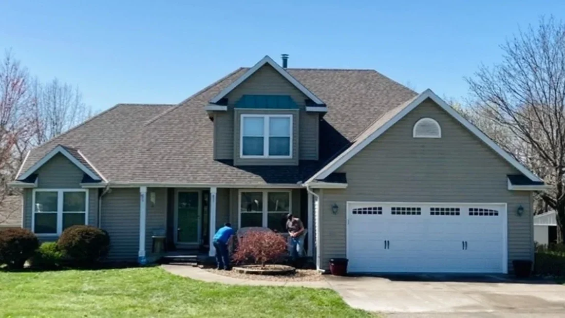 Two people working outside a two-story suburban house with a green lawn and a garage.