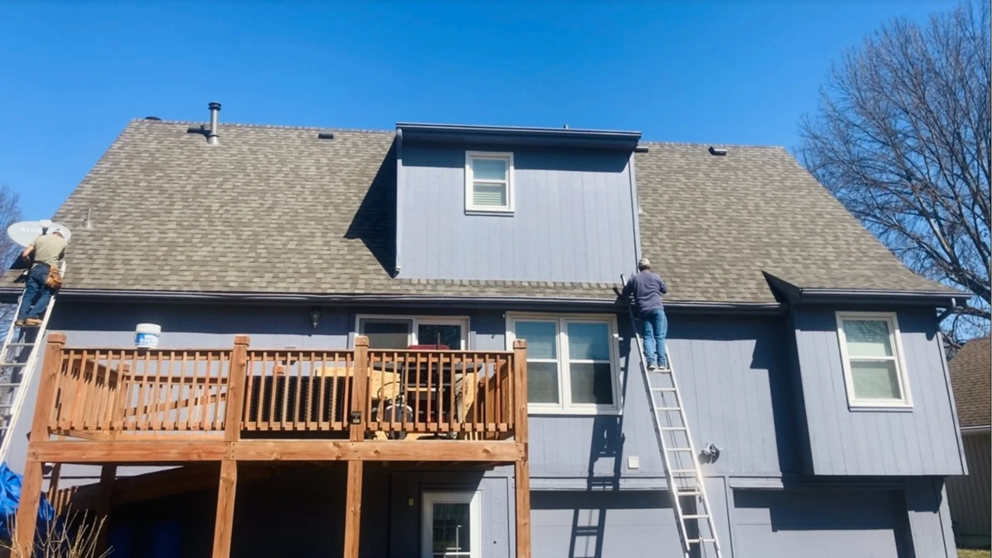 Two workers on ladders repairing the roof of a blue house with a balcony and windows, clear blue sky, and leafless trees in the background.