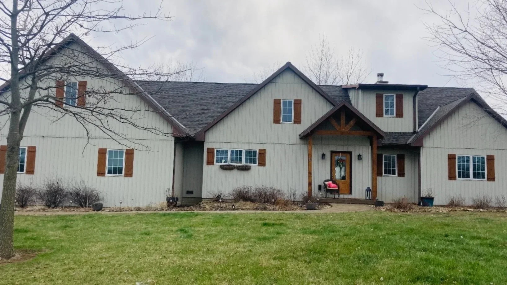 A large two-story house with a gray exterior and wooden accents. It has a front porch with a door decorated with a holiday wreath, and several windows with shutters. There is a well-maintained lawn and a few leafless trees, indicating it is winter or early spring.