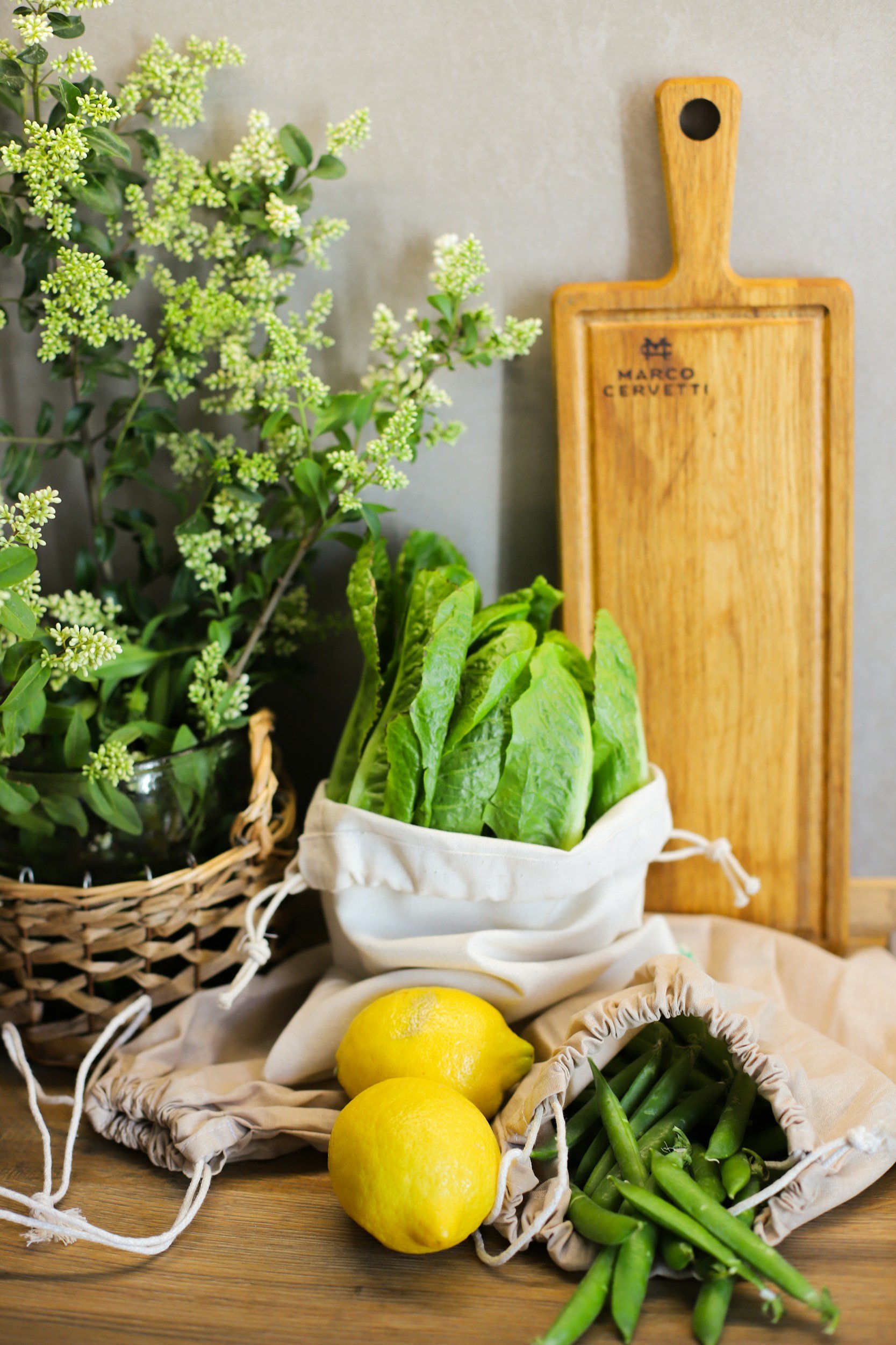 Fresh green lettuce, two lemons, and green beans in cloth bags on a wooden surface, with a cutting board and potted greenery in the background.