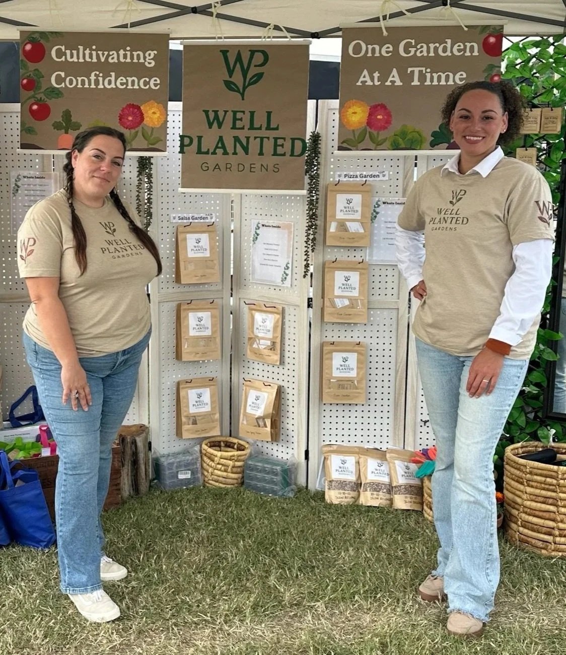 Two women standing in front of a display booth at a garden event, with signs that say 'Cultivating Confidence,' 'One Garden at a Time,' and 'Well Planted Gardens.' They are wearing matching beige T-shirts with the company's logo. The booth features various plant-related materials and informational flyers.