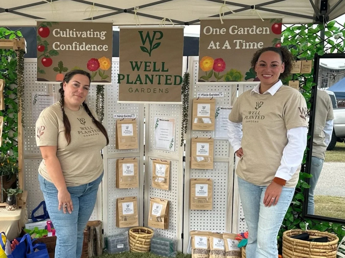 Two women standing at a booth displaying gardening products with signs that say "Cultivating Confidence" and "One Garden At A Time". The booth features a display labeled "Well Planted Gardens".