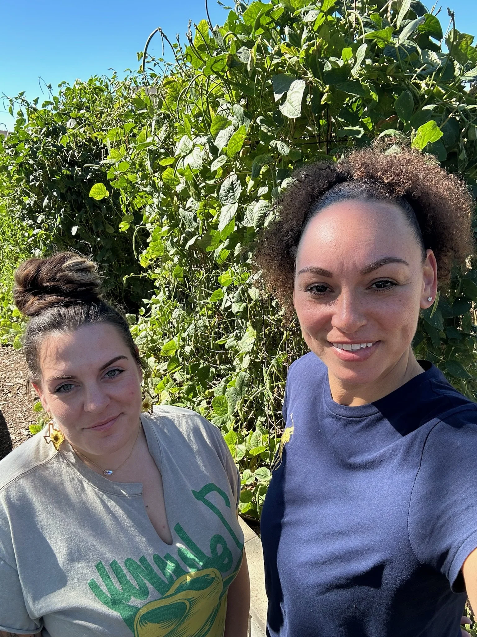Two women taking a selfie outdoors in front of lush green plants under a clear blue sky.