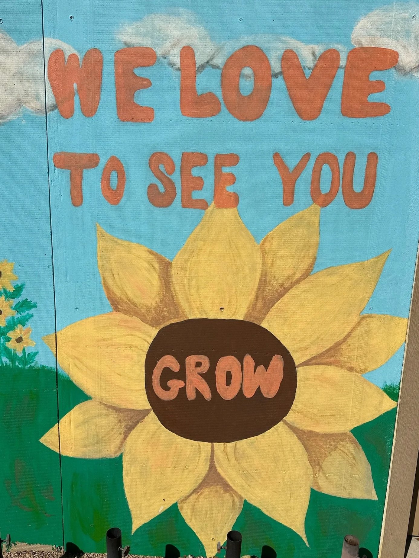 A painted mural with a bright blue sky, clouds, green grass, and a large sunflower with yellow petals and a brown center. The words "WE LOVE TO SEE YOU GROW" are written in large, orange letters across the top, with the word "GROW" inside the sunflower's center.
