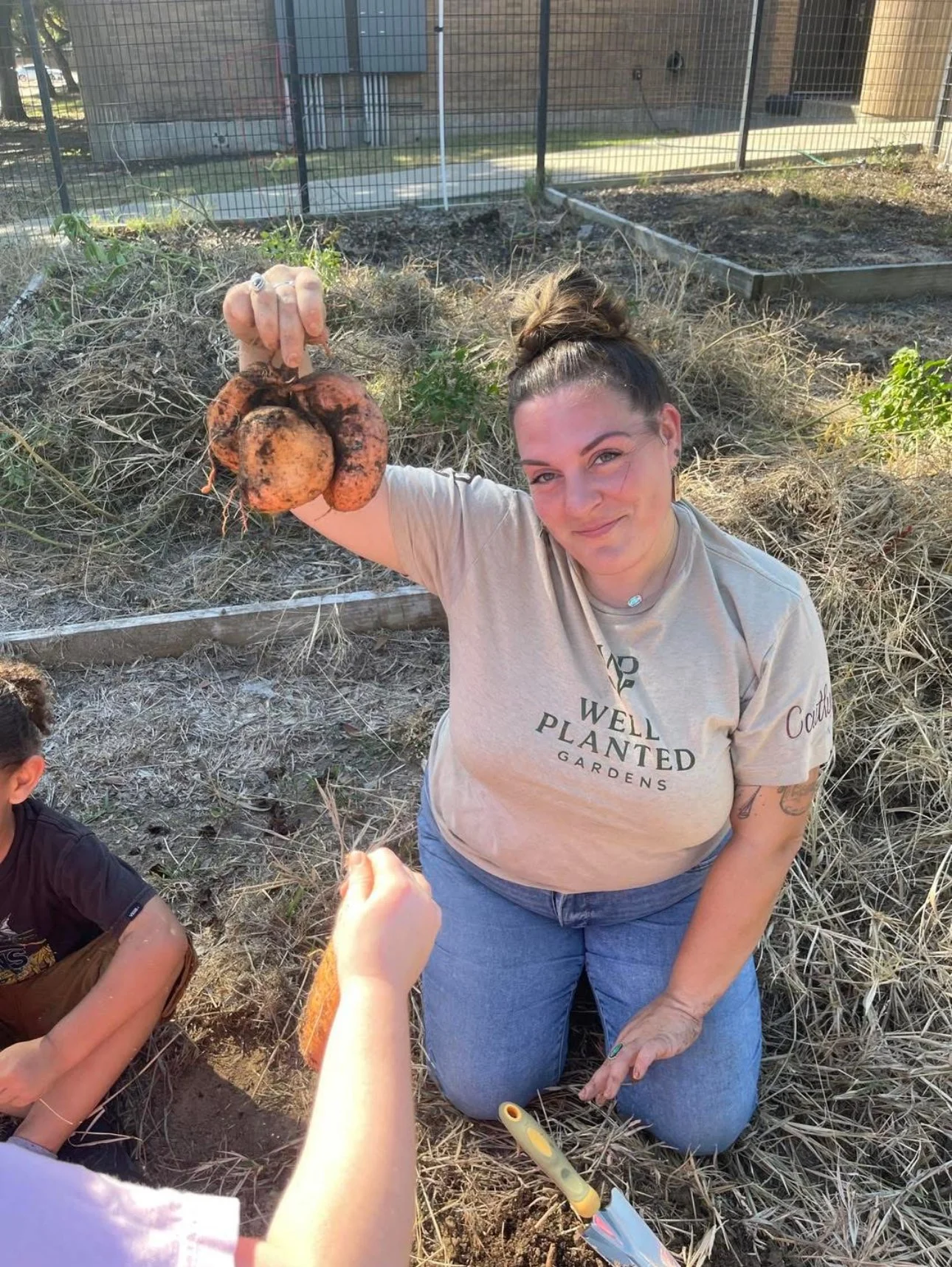 Woman kneeling in a garden holding freshly harvested carrots covered in dirt, with a child's hand offering a carrot in the foreground.