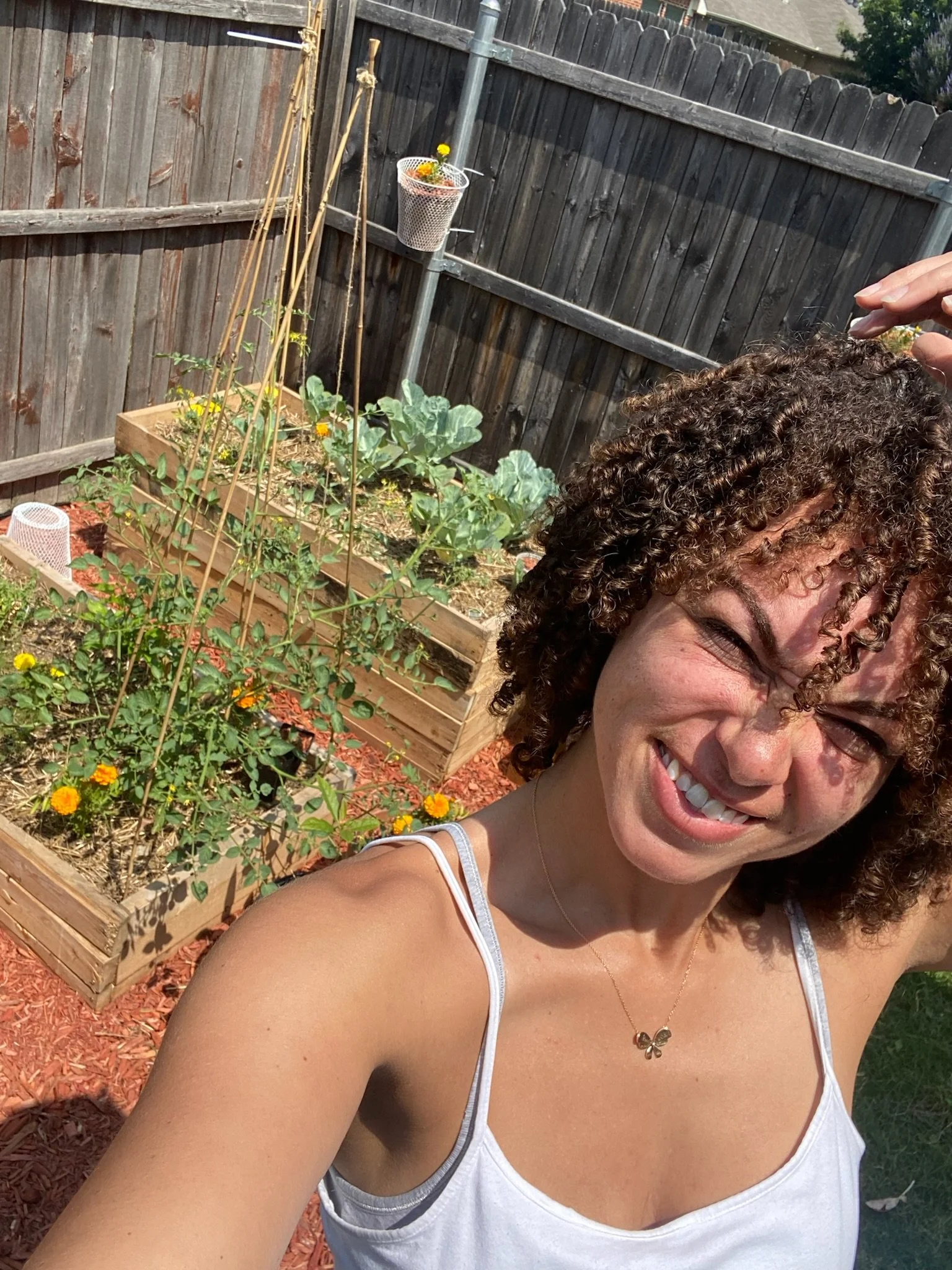 A woman with curly hair taking a selfie in a garden with raised wooden beds, plants, and a wooden fence.