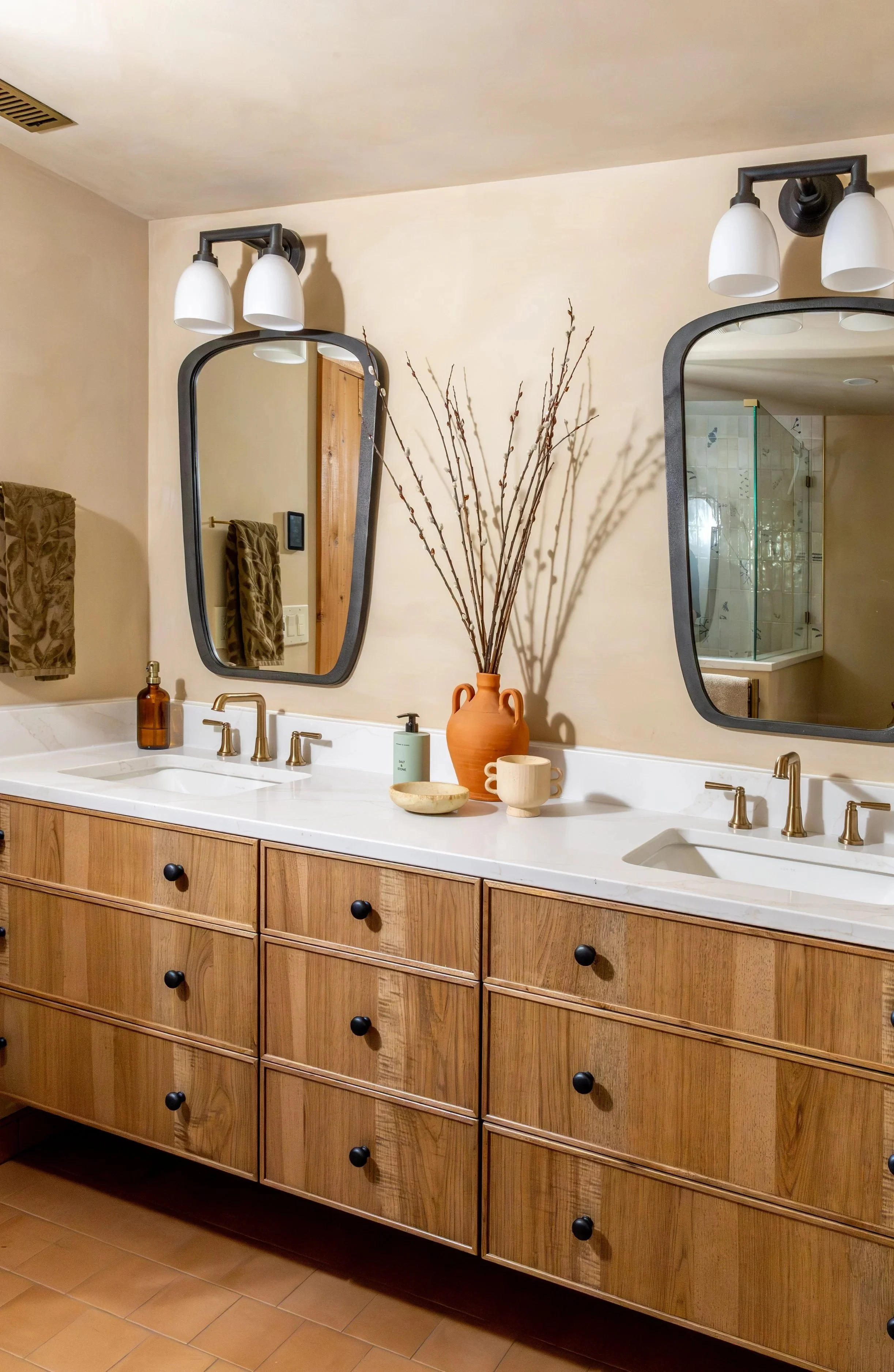 A bathroom vanity with a wooden cabinet, white countertop, and two sinks. Decor includes a brown soap dispenser, a white mug, a green hand soap bottle, a ceramic bowl, and a large terra cotta vase with bare branches. Two mirrors with black frames and matching black light fixtures are mounted above the sinks. The wall is painted beige, with towel rings holding matching patterned towels and a wooden door visible in the reflection. The bathroom features tiled flooring.