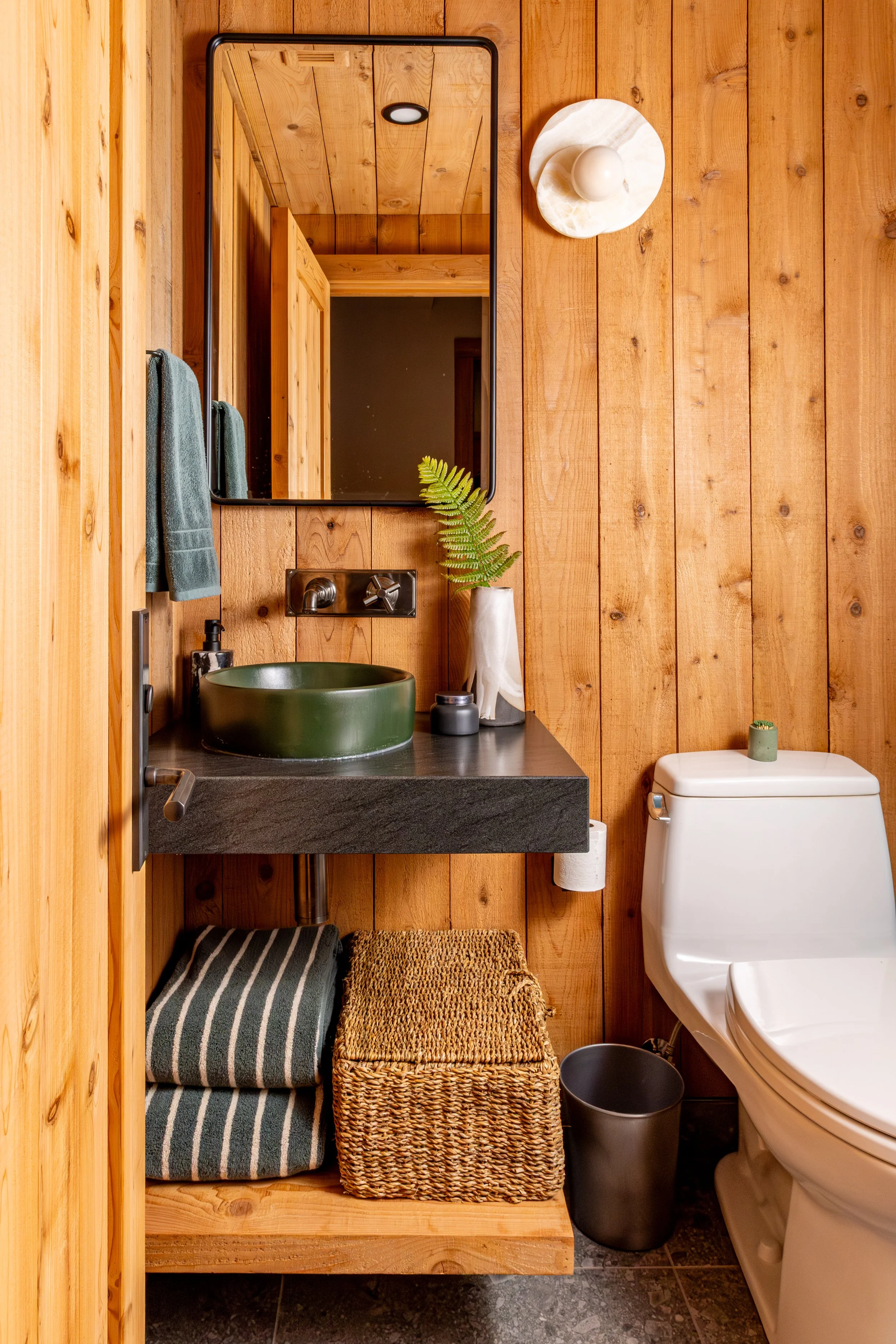 A small rustic bathroom with wood-paneled walls, a mirror, a green vessel sink, a faucet, a white soap dispenser, a small green plant, a towel, a woven basket, a grey trash can, a beige toilet, and a round marble decorative item on the wall.