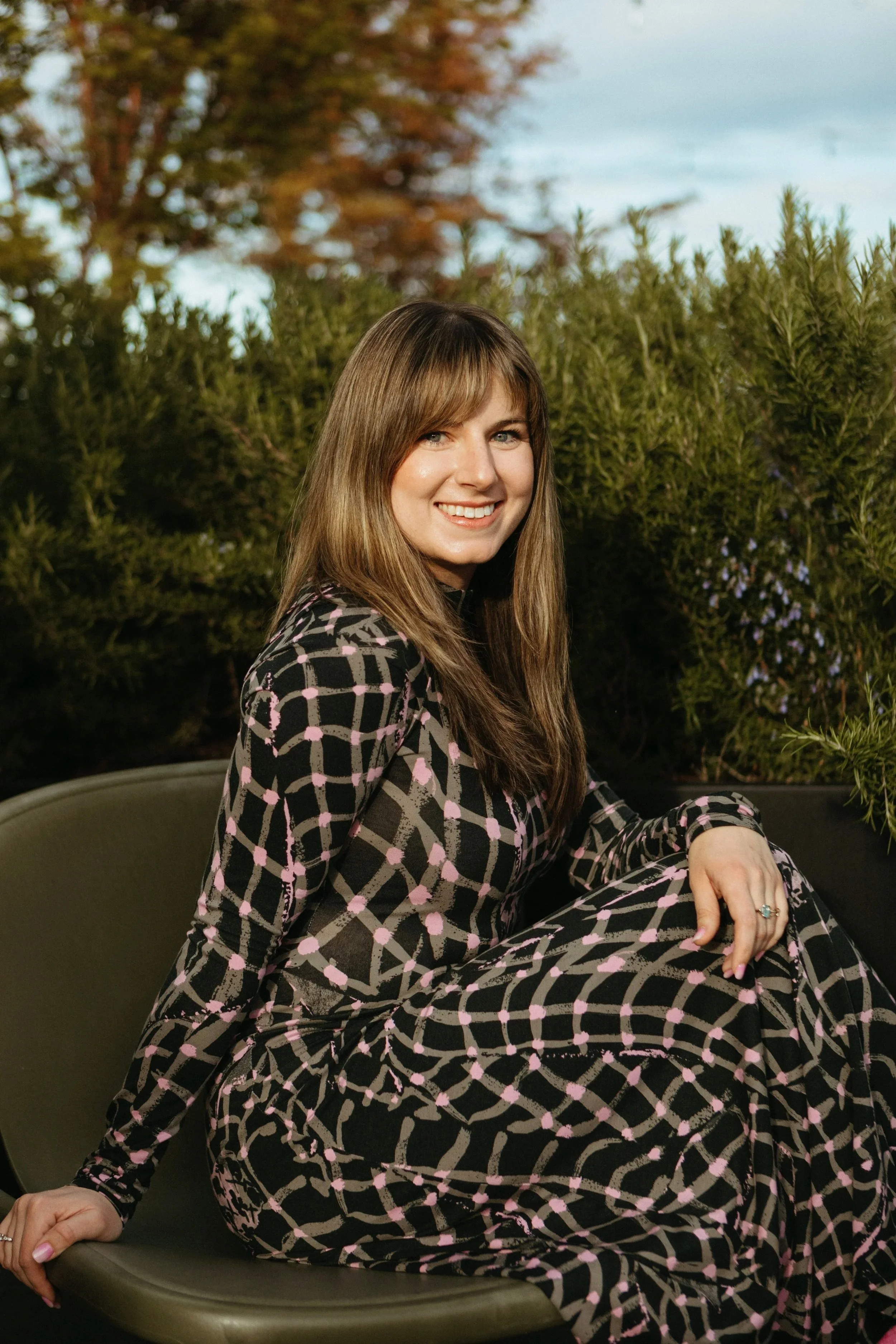 A smiling woman with long, light brown hair and blue eyes sitting outdoors. She's wearing a black dress with a pink and brown geometric pattern. The background consists of green shrubs and a partly cloudy sky.