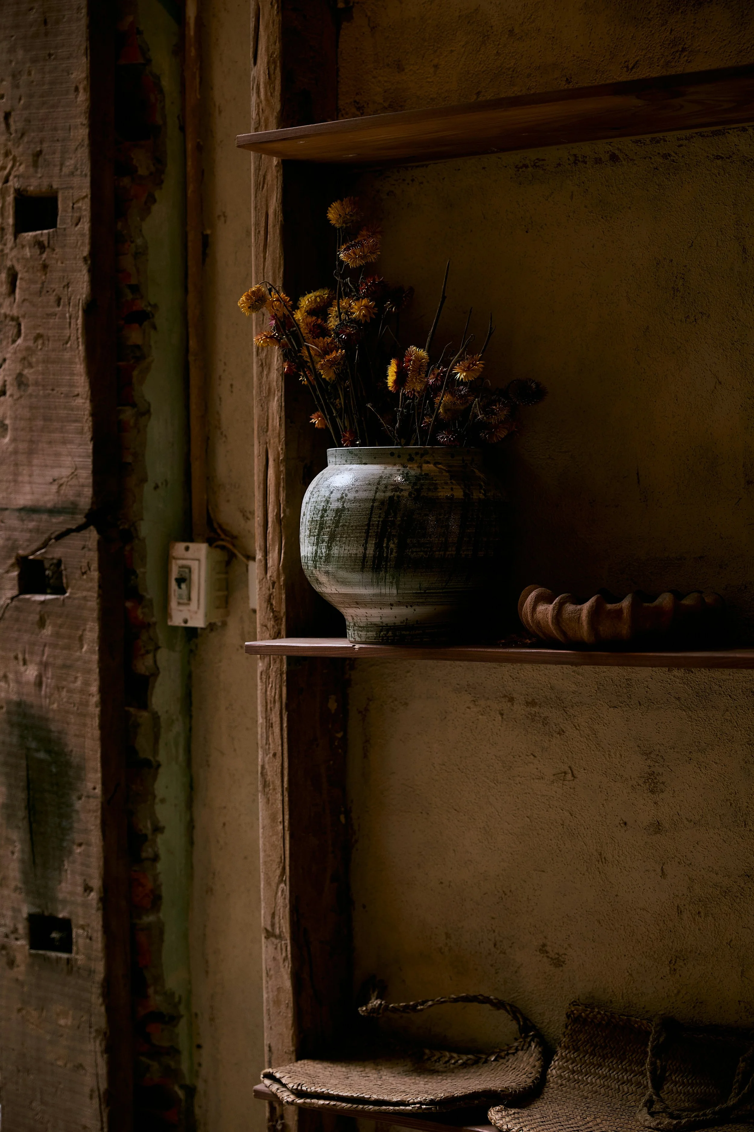 A rustic interior with a large ceramic vase filled with dried yellow and orange flowers on a wooden shelf, next to two woven chairs with rope armrests.