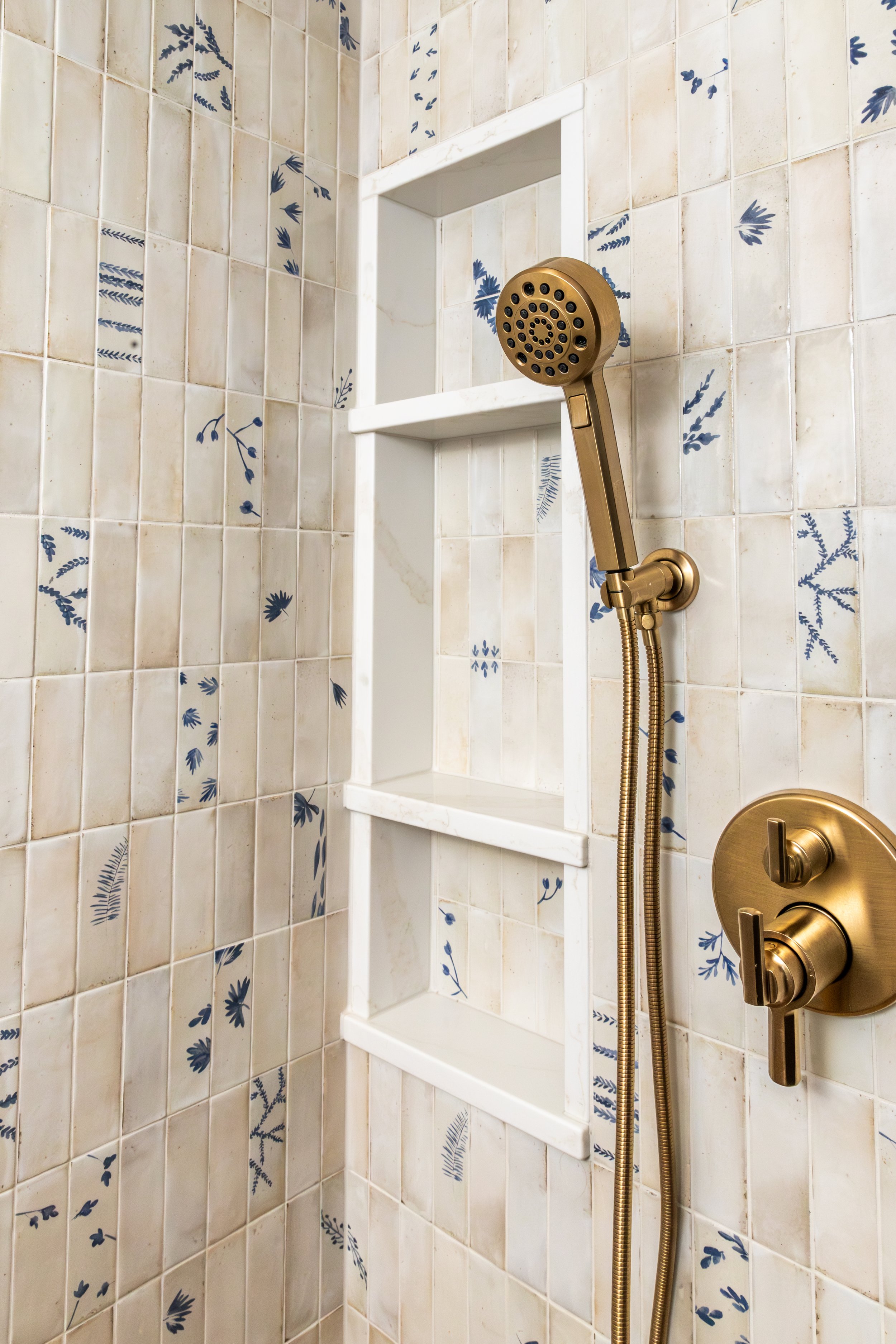 Shower with beige and blue floral tiles, built-in white shelves, and gold fixtures including a showerhead and control handles.