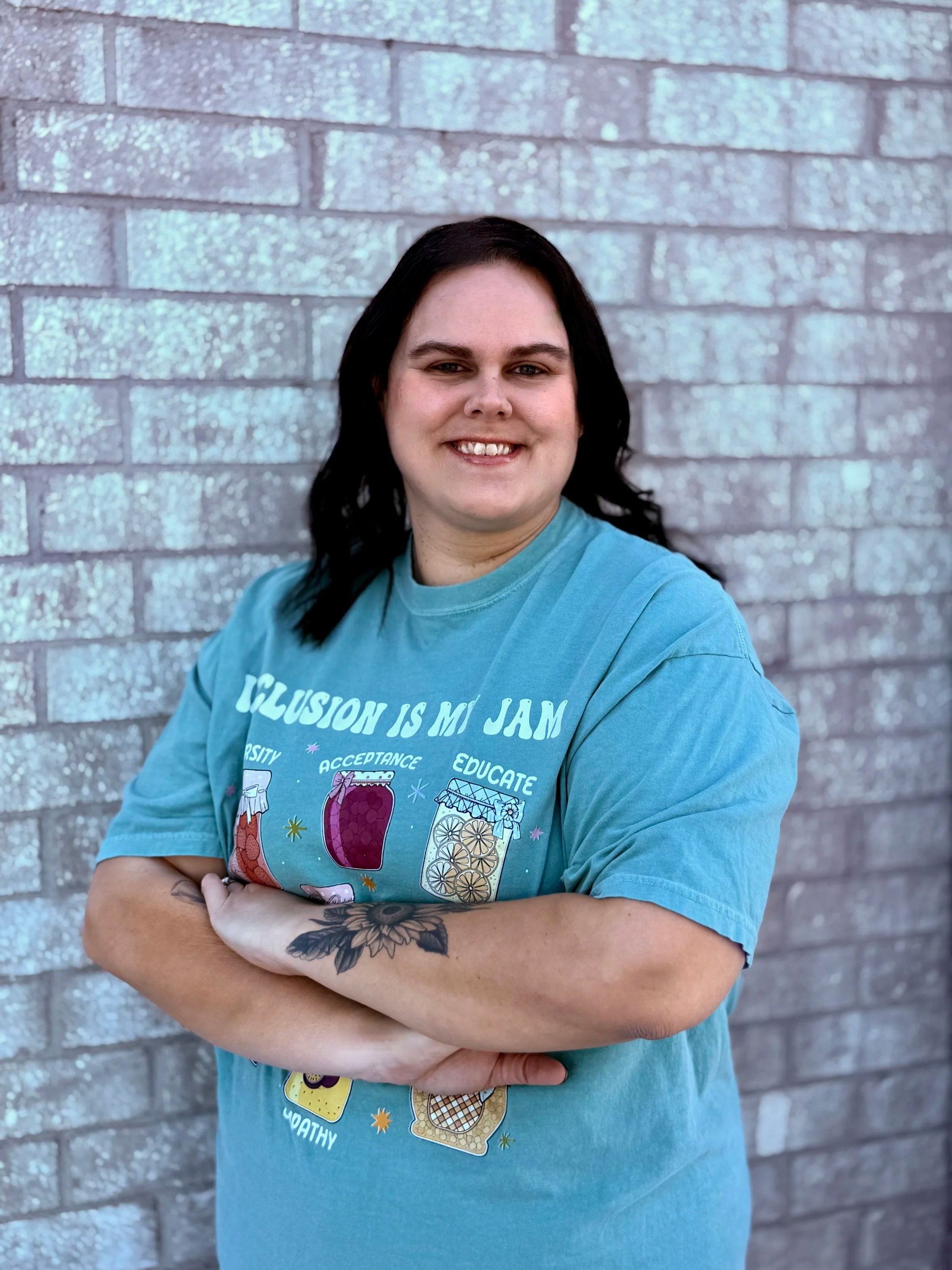 A woman with dark hair and a tattoo on her forearm smiling and standing with arms crossed against a gray brick wall, wearing a blue T-shirt with colorful graphic designs and text.