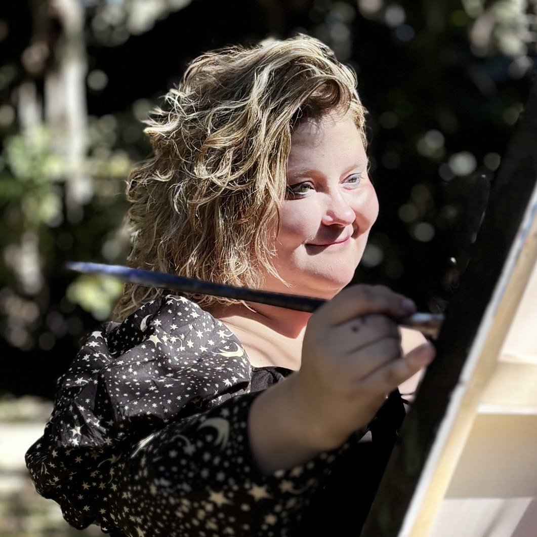 A woman with curly blonde hair painting outdoors on a canvas.