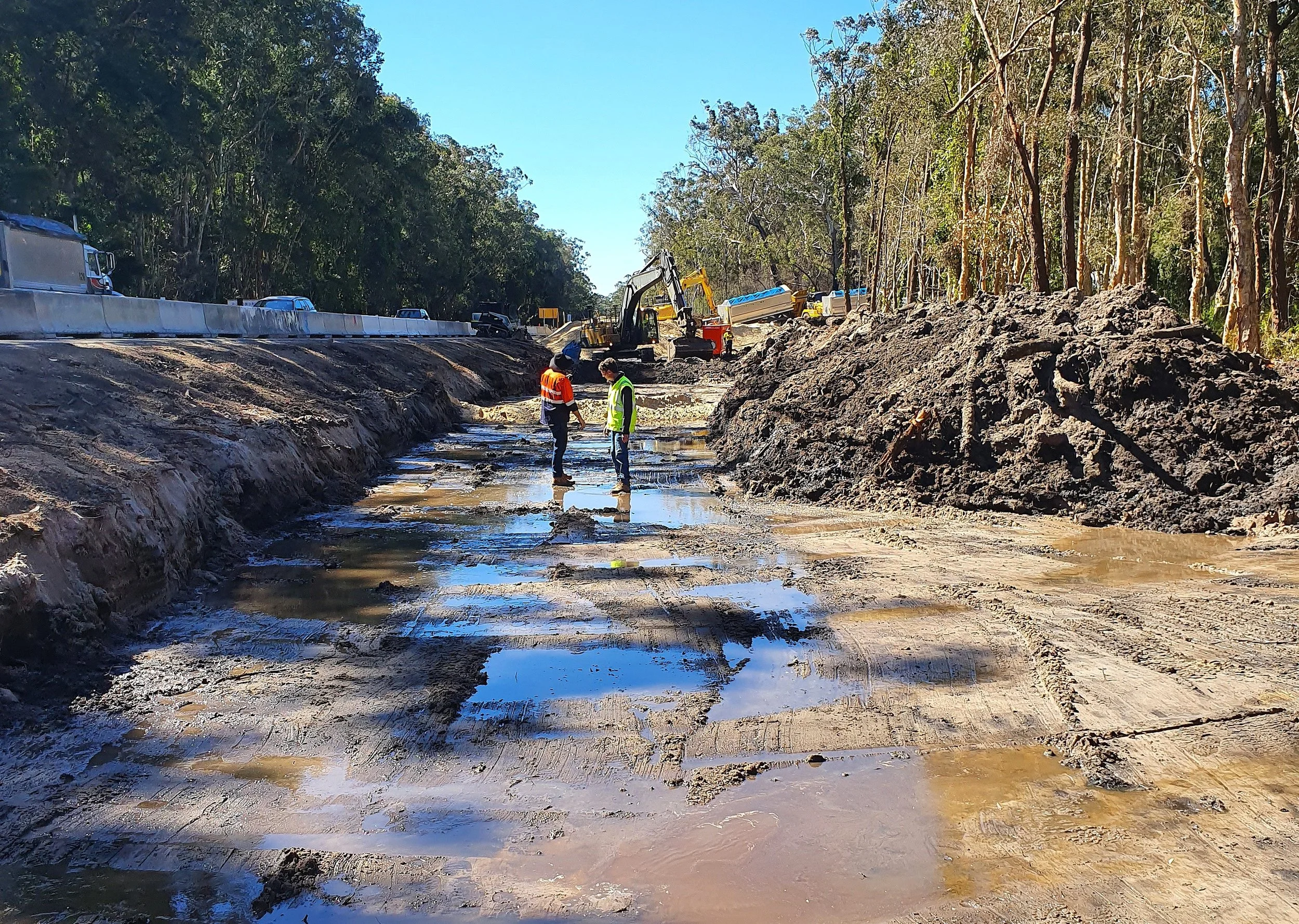 Construction workers inspecting a muddy road under construction, with dirt and machinery on the site, and cars traveling on the nearby highway.