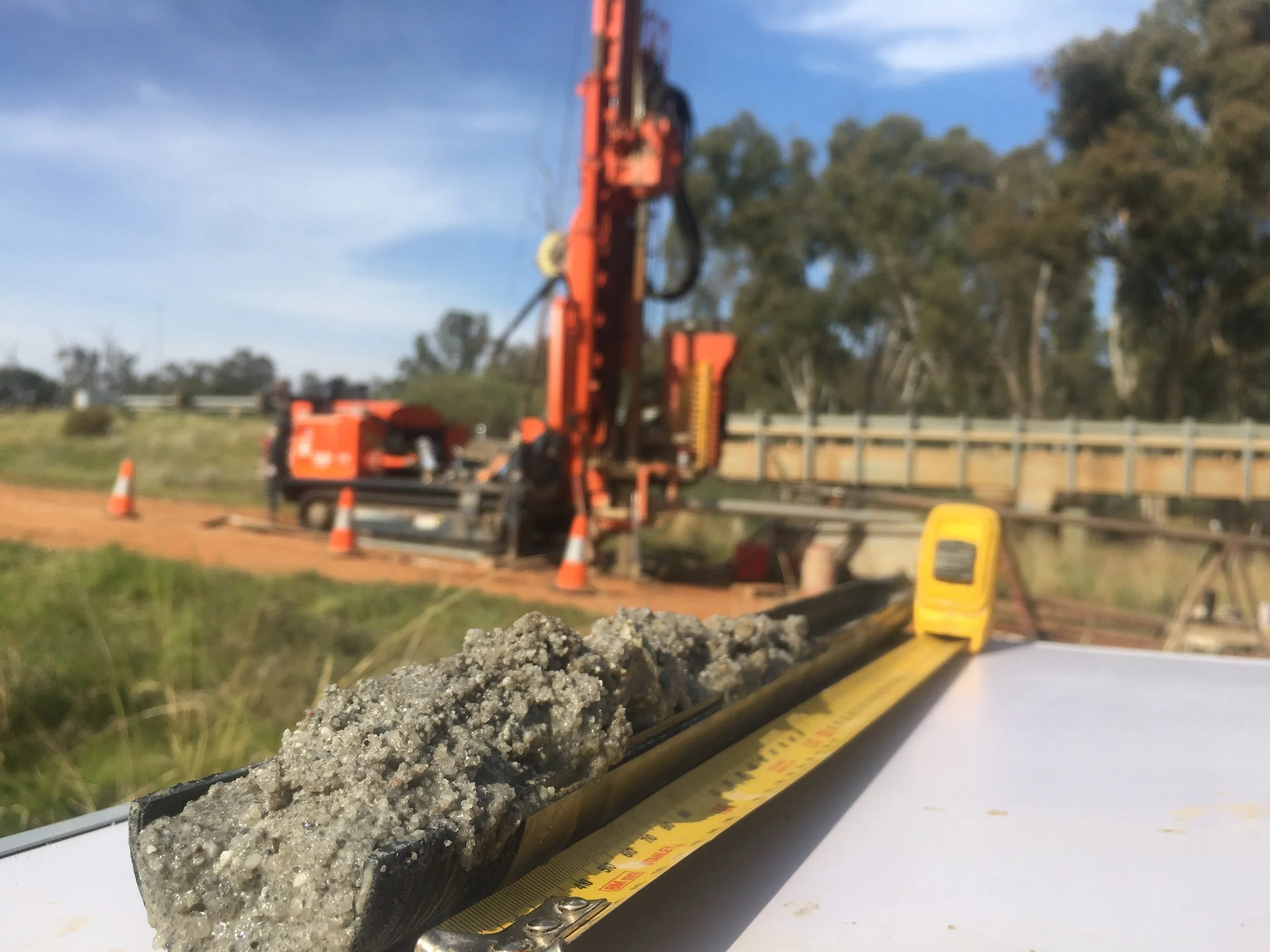 A construction site with a focus on a measuring tool and concrete sample in the foreground, and construction machinery and safety cones in the background.