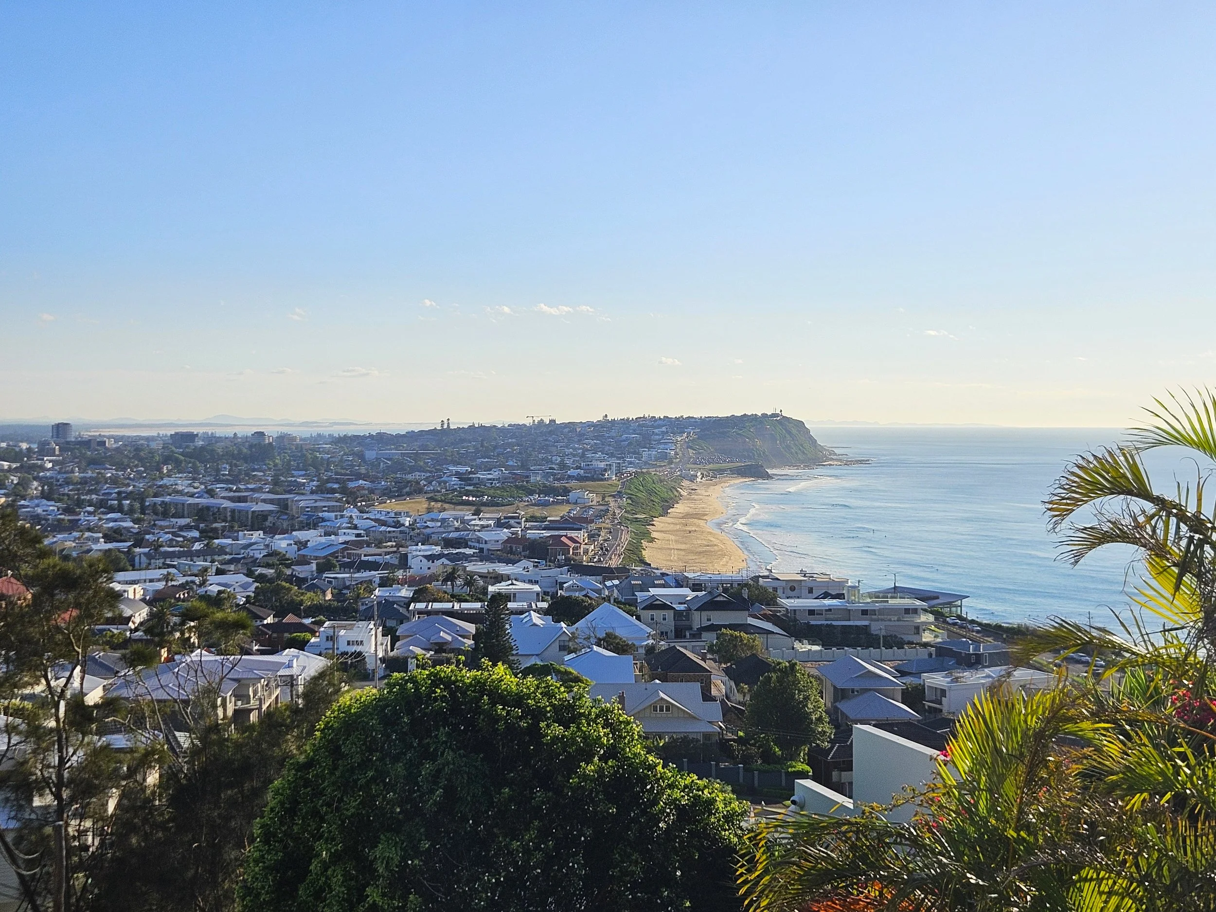 A coastal cityscape with numerous houses, a beach, and a cliff in the distance on a sunny day.