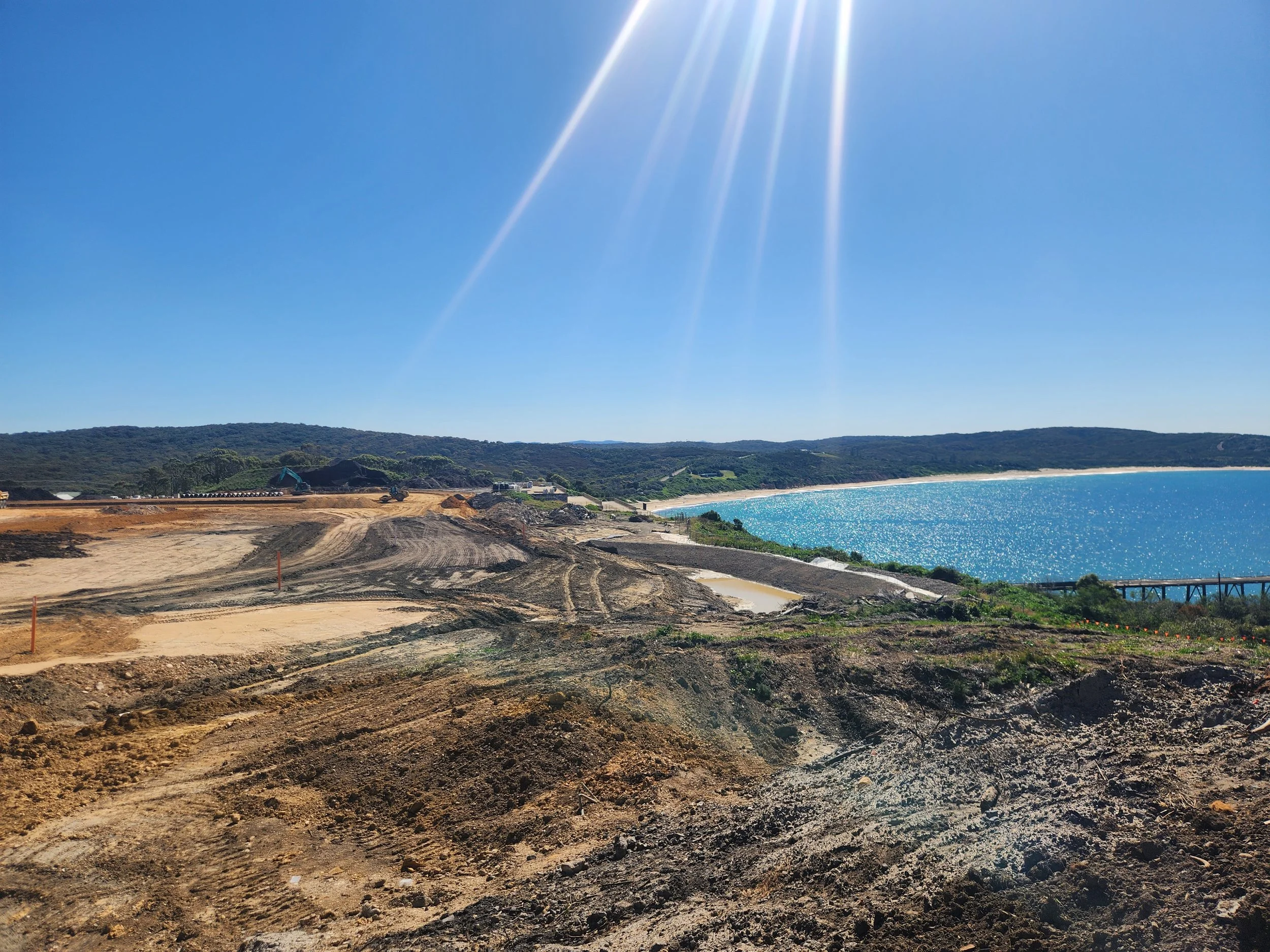 Construction site near the beach with dirt mounds, heavy machinery, and a train track, under a clear blue sky with sunlight.