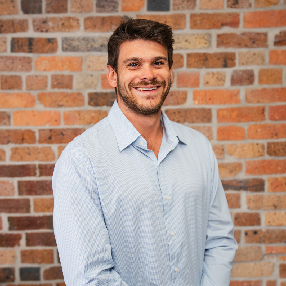Black and white photo of a young man with short, styled hair, smiling slightly, wearing a dark shirt, with a brick wall in the background.
