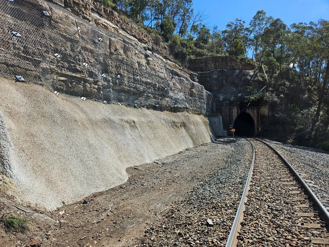 Railway tracks leading into a tunnel through a rocky hillside under a clear blue sky surrounded by trees.