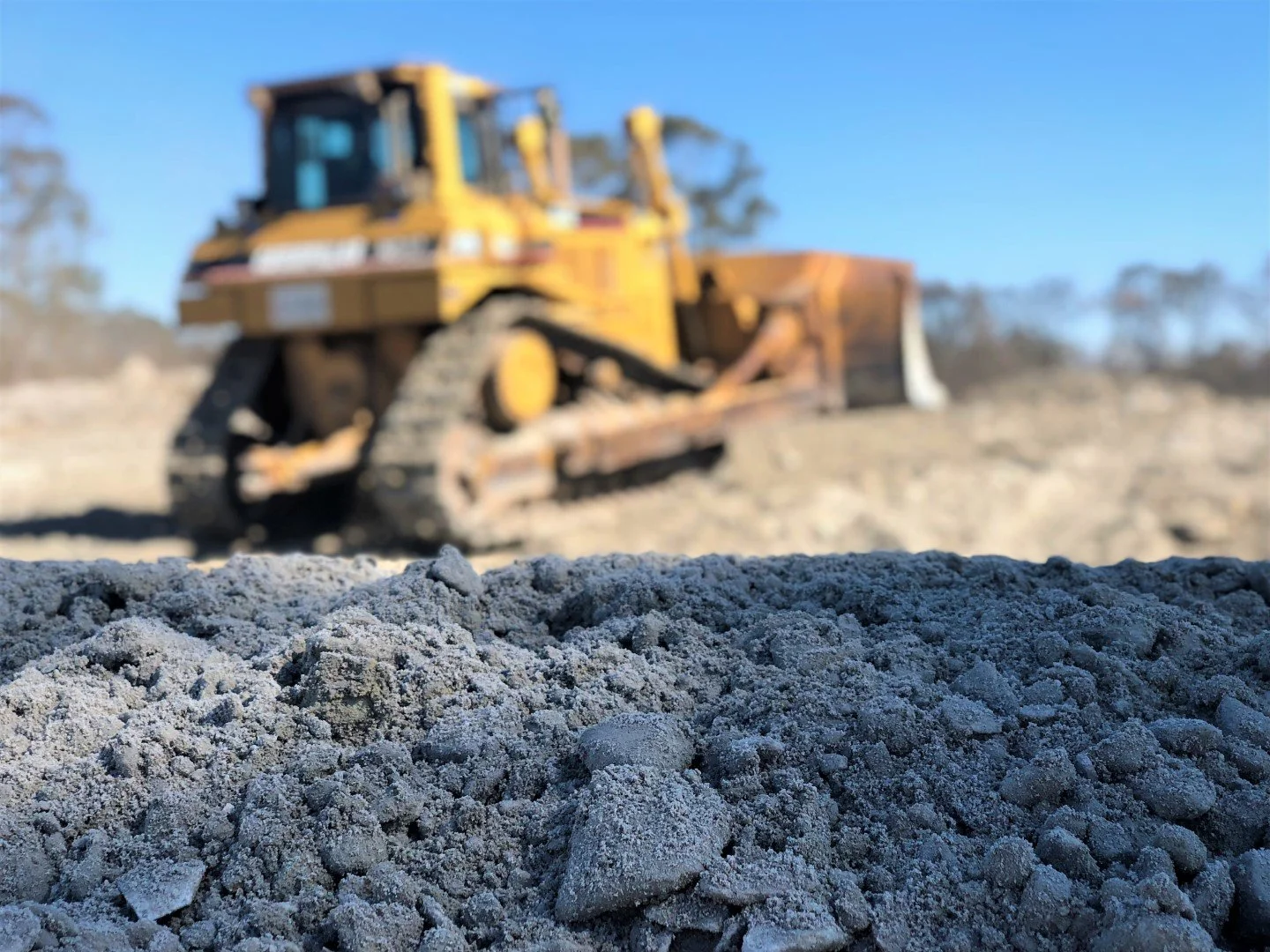 Close-up of a mound of dirt or gravel with a blurred yellow bulldozer in the background under a clear blue sky.