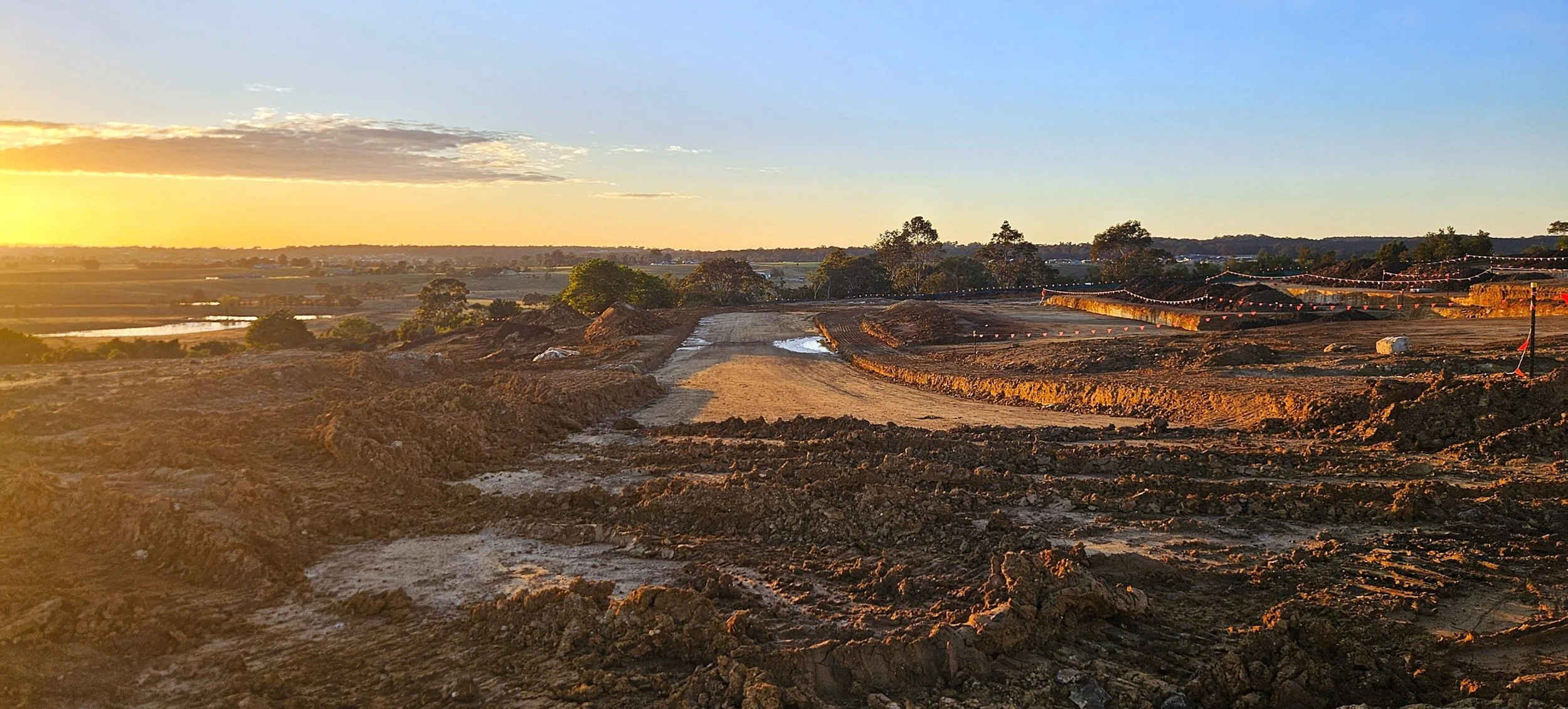 Construction site with dirt tracks and equipment, gathered at sunset or sunrise, with trees and landscape in the background.