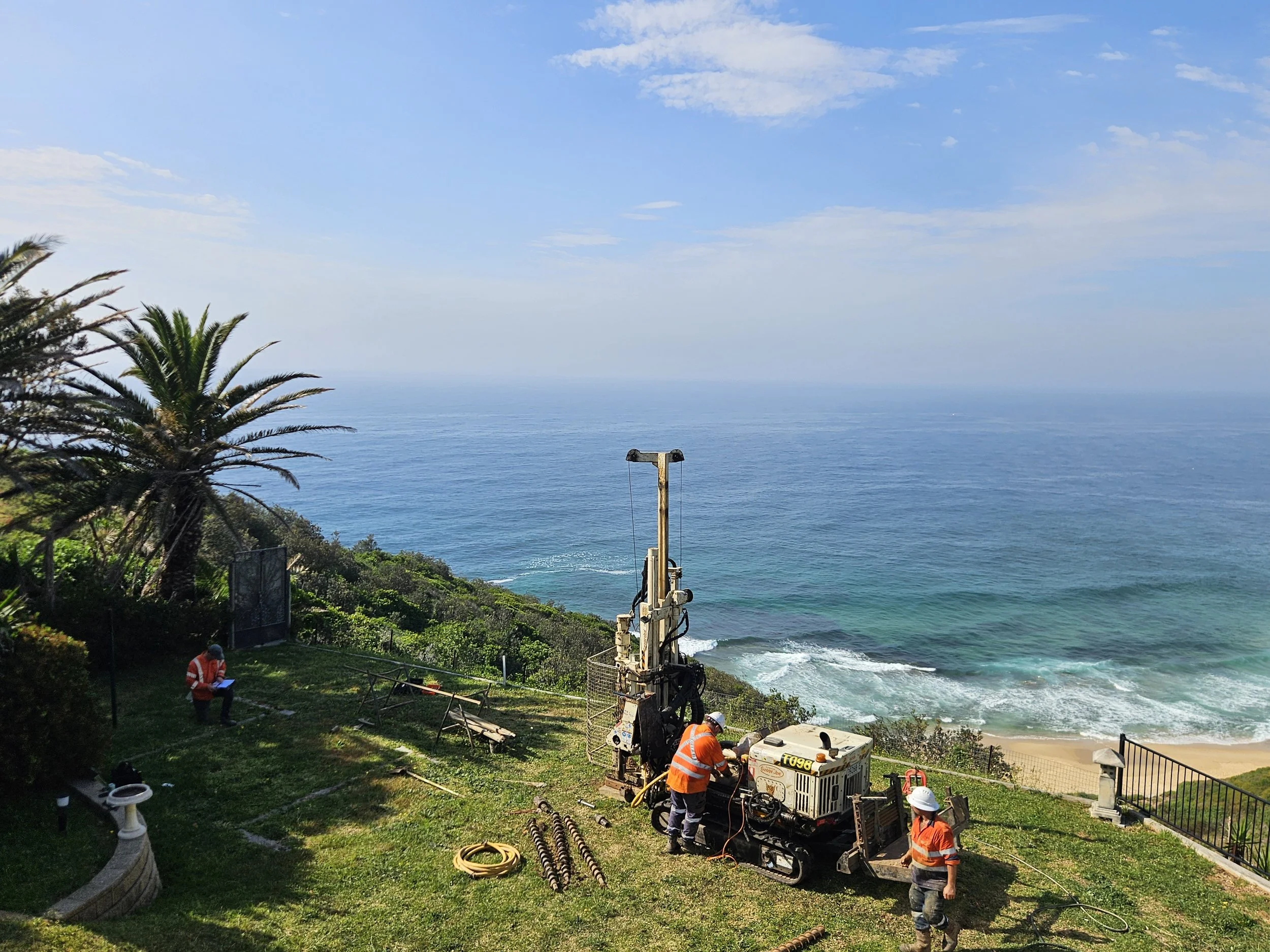 Construction workers operating equipment near a coastal cliff with palm trees, a sandy beach, and the ocean in the background.
