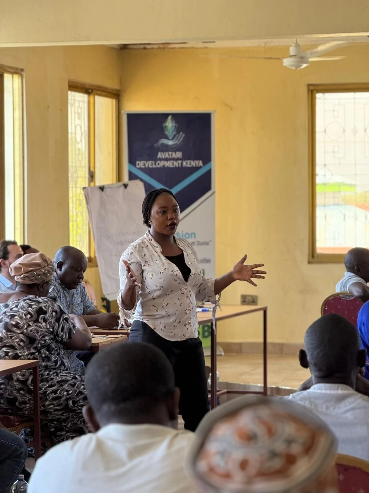 A woman standing and speaking to an audience in a room with yellow walls and windows, with a banner in the background that reads "AVATARI DEVELOPMENT KENYA."