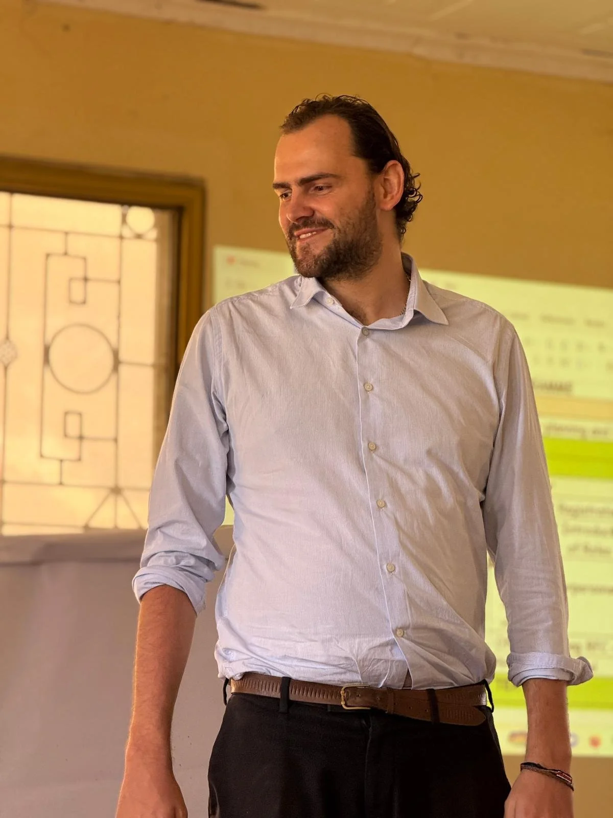 A man with dark, curly hair and a beard, wearing a white button-down shirt with sleeves rolled up, standing in a room with warm lighting and a window with stained glass design in the background.