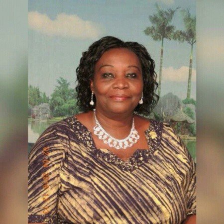 A woman with short, curly black hair wearing a patterned dress, a pearl necklace, and pearl earrings, standing outdoors with a background of palm trees and a scenic landscape.