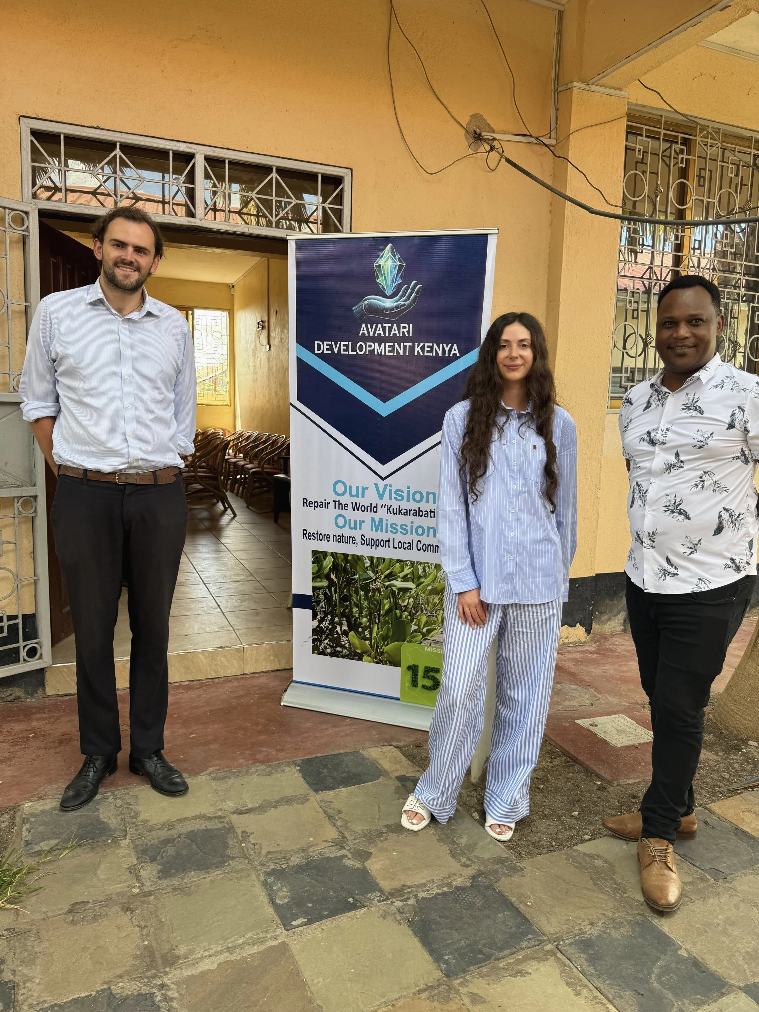 Three people standing outdoors in front of a banner that reads "Avatari Development Kenya". The banner also displays their vision and mission statements. The two men are wearing button-up shirts and the woman is in striped pajamas. They are smiling a