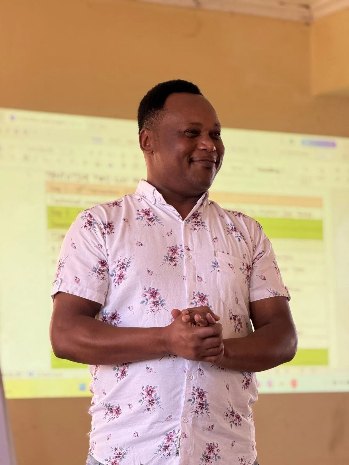 A man with short dark hair and a dark skin tone, wearing a light pink, short-sleeved shirt with a floral pattern, is standing with his hands clasped, smiling in front of a blurred presentation or whiteboard.