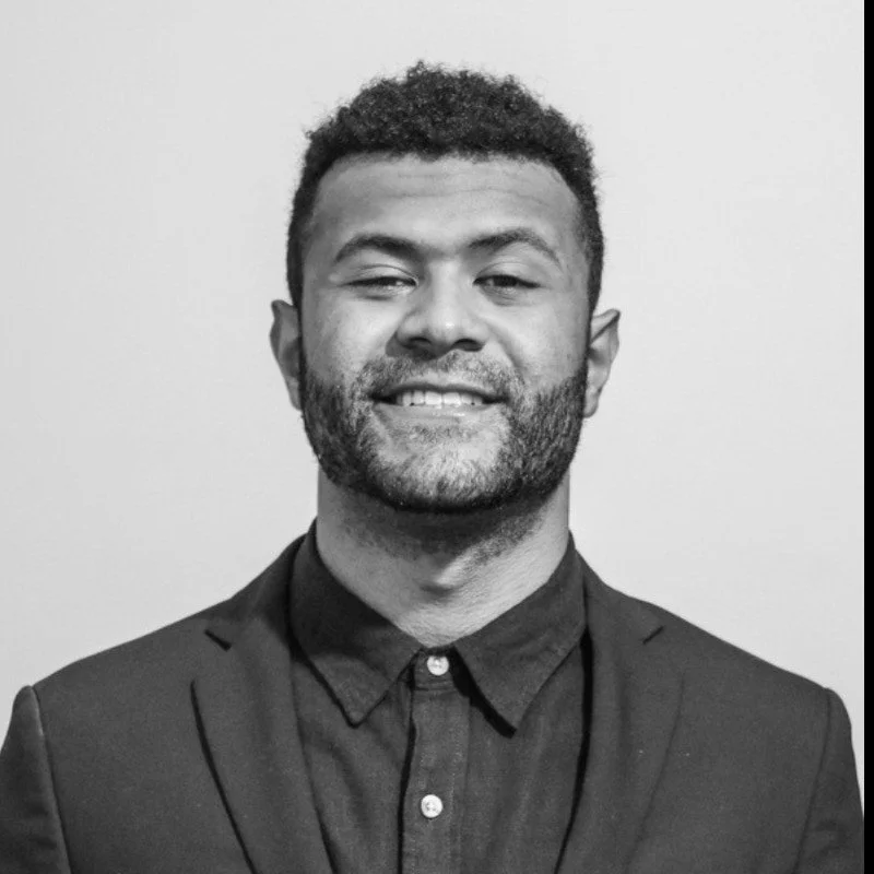 A man with dark curly hair and a beard, wearing a dark blazer and a button-up shirt, smiling at the camera against a plain background.