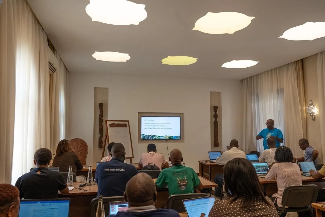 A man in a blue shirt giving a presentation to an audience seated at tables using laptops in a conference room with light curtains and ceiling lights.