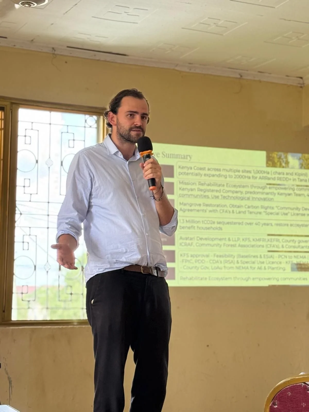 A man giving a presentation in front of a slide that contains text about mangrove restoration, with a window behind him showing a decorative iron grille and trees outside.