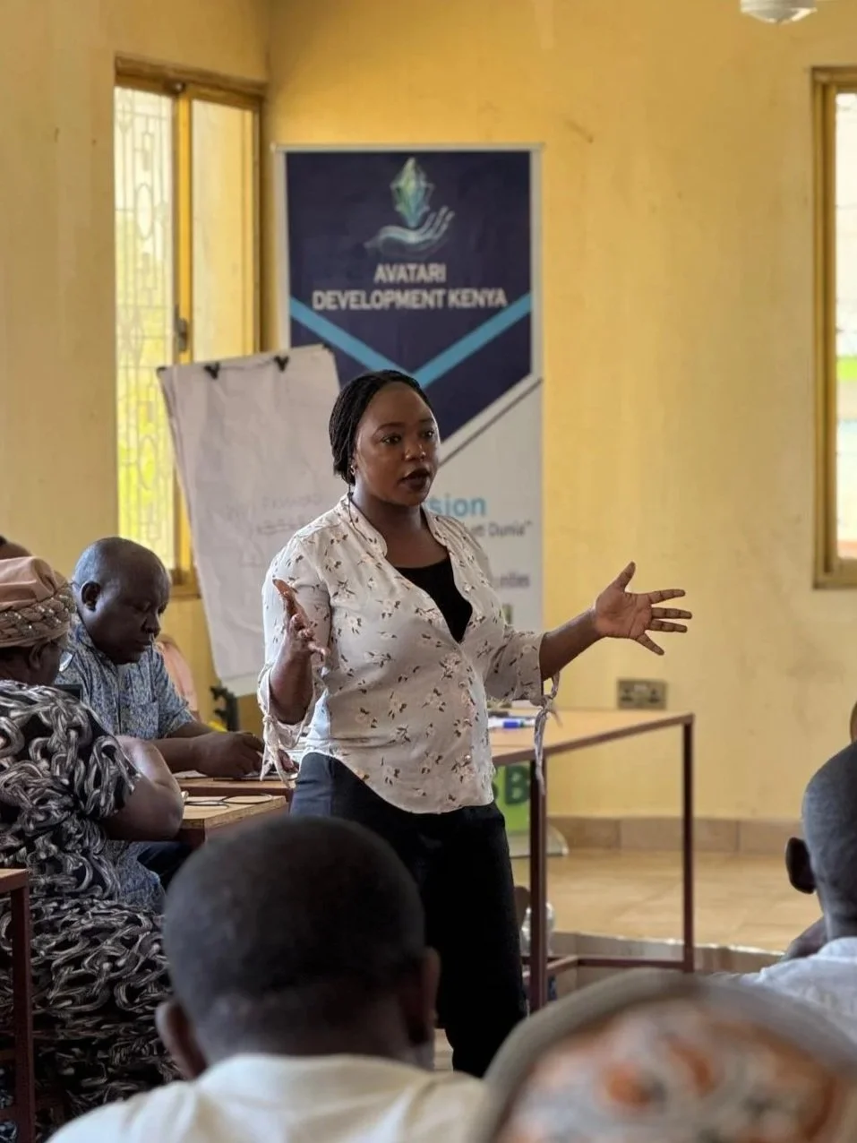 A woman giving a presentation in a room with several seated attendees. There is a banner in the background that reads 'Avatar Development Kenya.' The woman is gesturing with her hands while speaking.