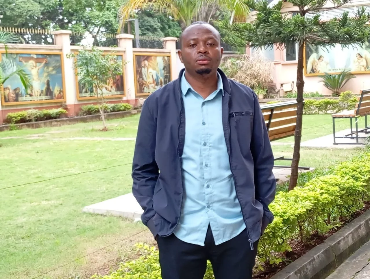 A man standing outdoors in a park area with benches and greenery, including trees and bushes, with religious paintings on a fence in the background.