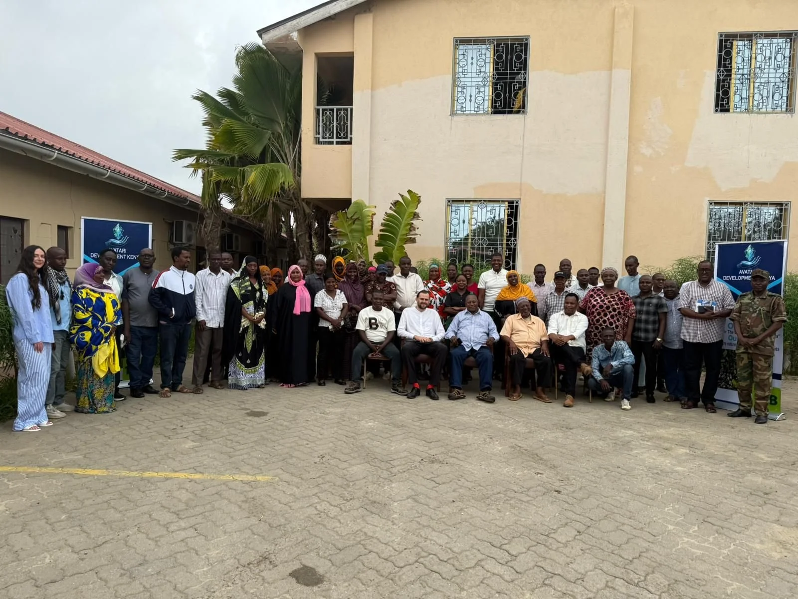 Group of diverse men and women gathered outdoors in front of a beige building with windows, facilitating a community or organizational event, with banners visible on either side. community meet up with team