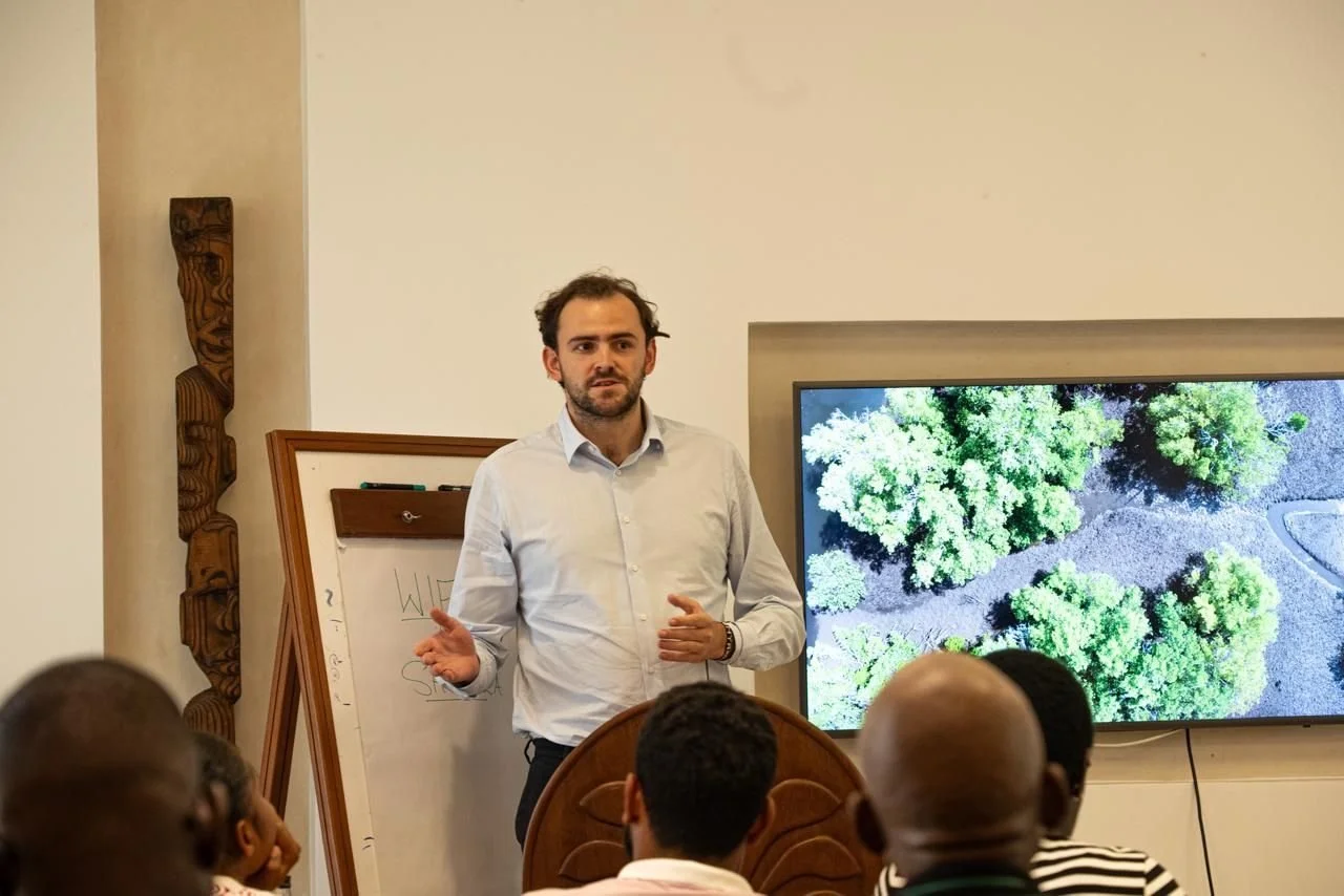 A man in a light gray shirt giving a presentation to a group of people in a conference room, with a large screen displaying an aerial view of green trees and a whiteboard behind him.