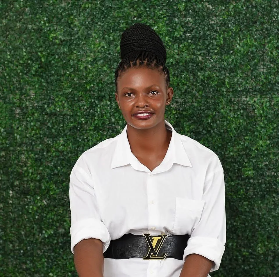 A woman with braided hair in a bun, wearing a white shirt and a black belt with a gold logo, standing in front of a green leafy background.