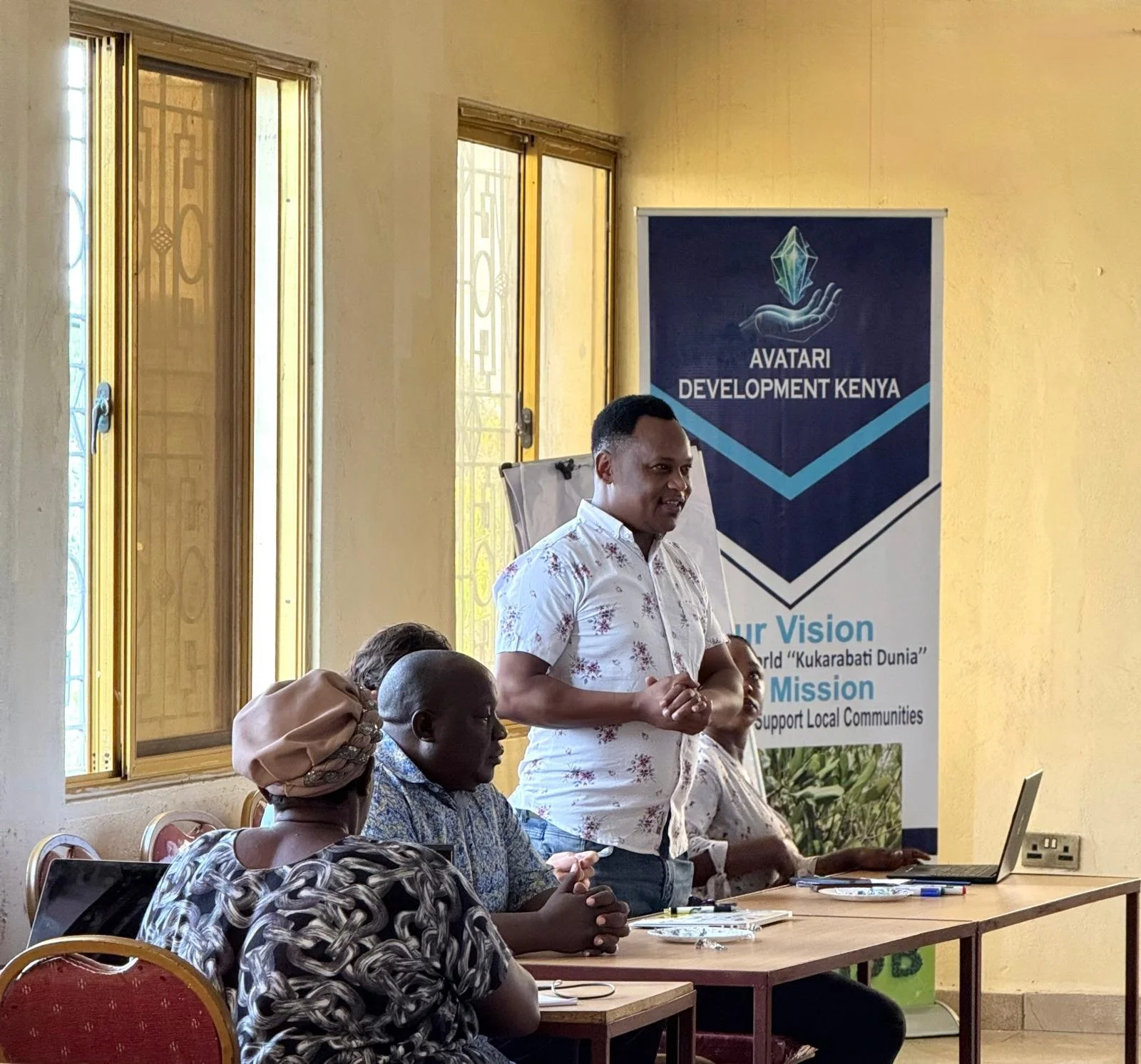 A man standing at a table with four seated adults, speaking during a meeting or presentation in a room with yellow walls and windows. A banner in the background reads "AVATARI DEVELOPMENT KENYA" with the slogans "Our Vision: A Kukarabati Dunia" and "