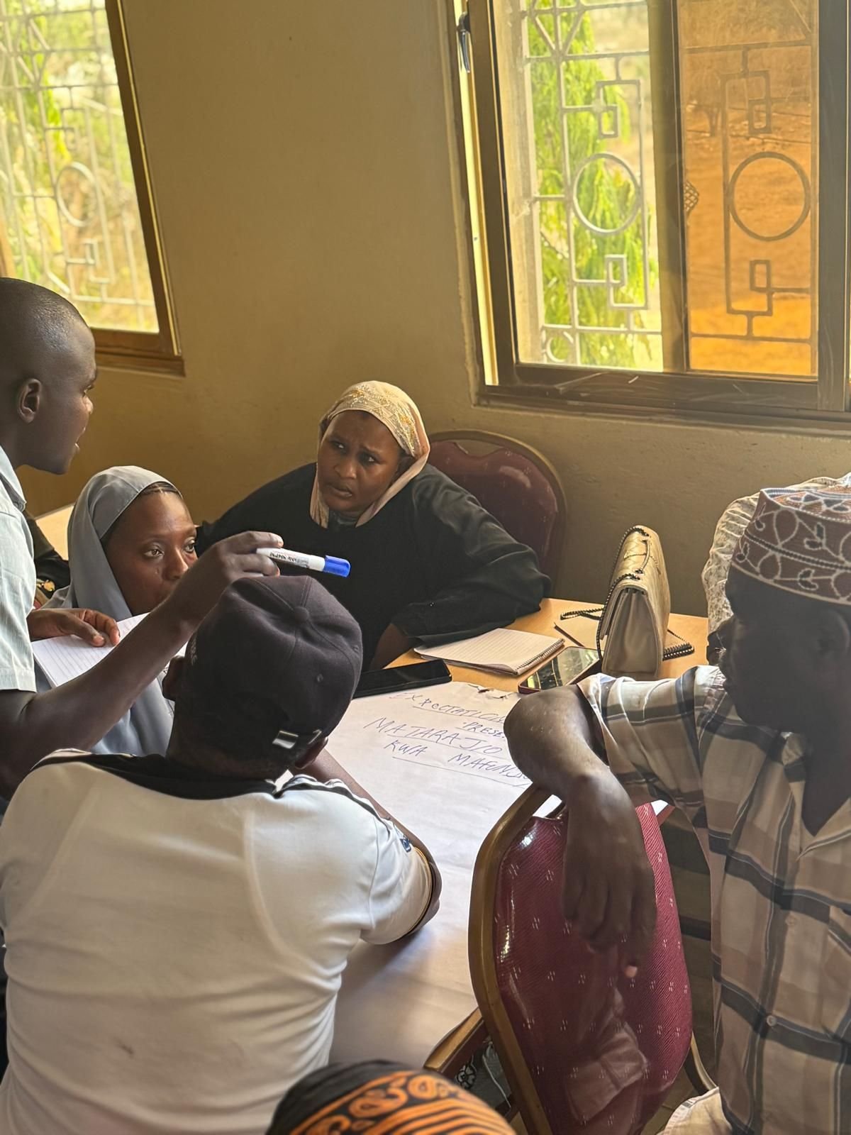 A group of people engaged in a discussion around a table, with notebooks, papers, and a whiteboard paper on the table, in a room with open windows and natural light.