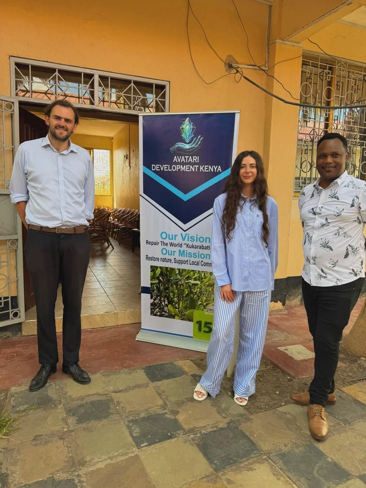 Three people standing outside in front of a banner for Avatari Development Kenya. The banner displays their vision and mission statements. The woman in the middle is wearing light blue pajamas, and the men on either side are dressed in dress shirts and dark pants.