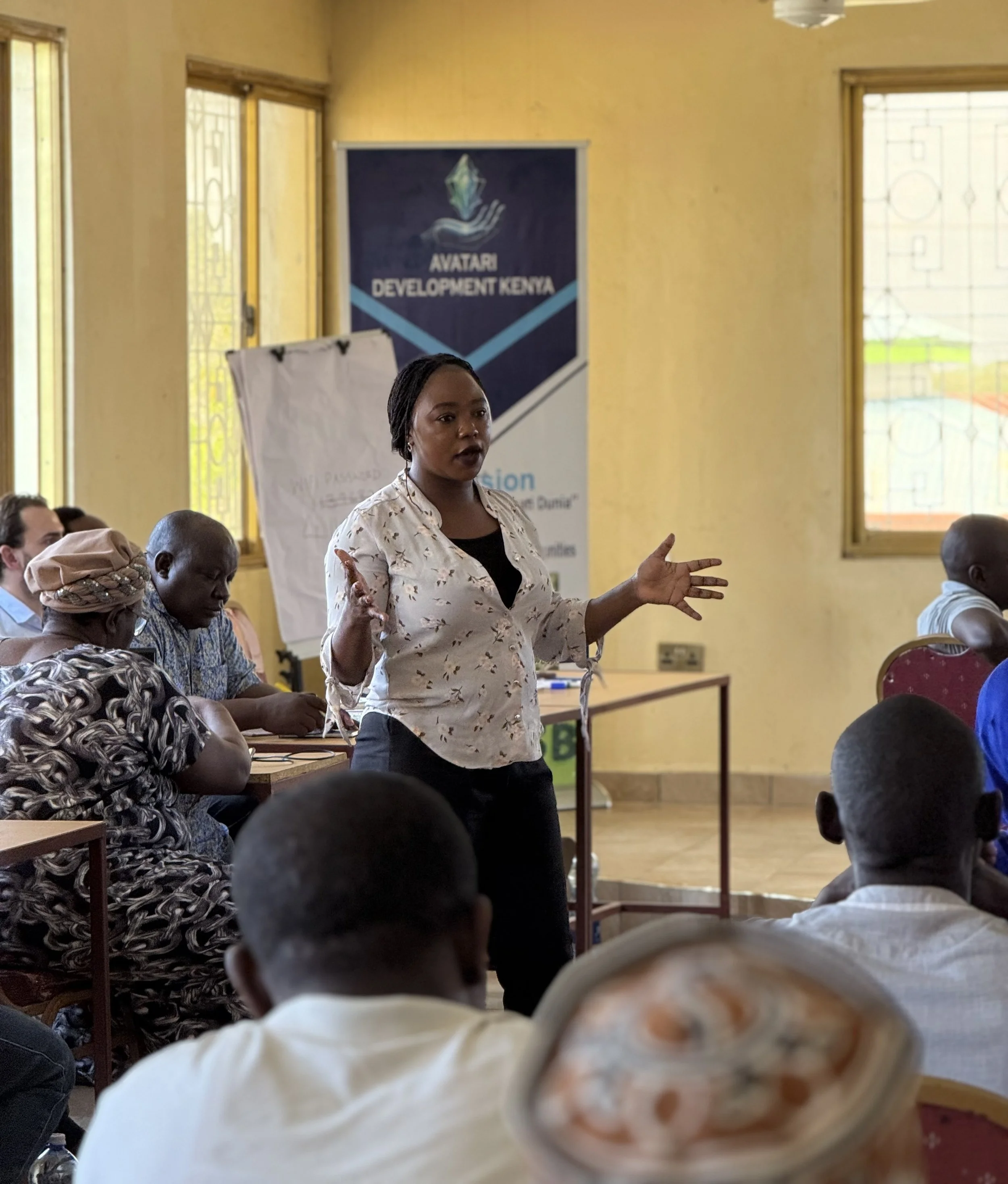 A woman standing and speaking to a group of people in a brightly lit room with yellow walls, large windows, and a banner that reads 'Avatar Development Kenya' in the background.