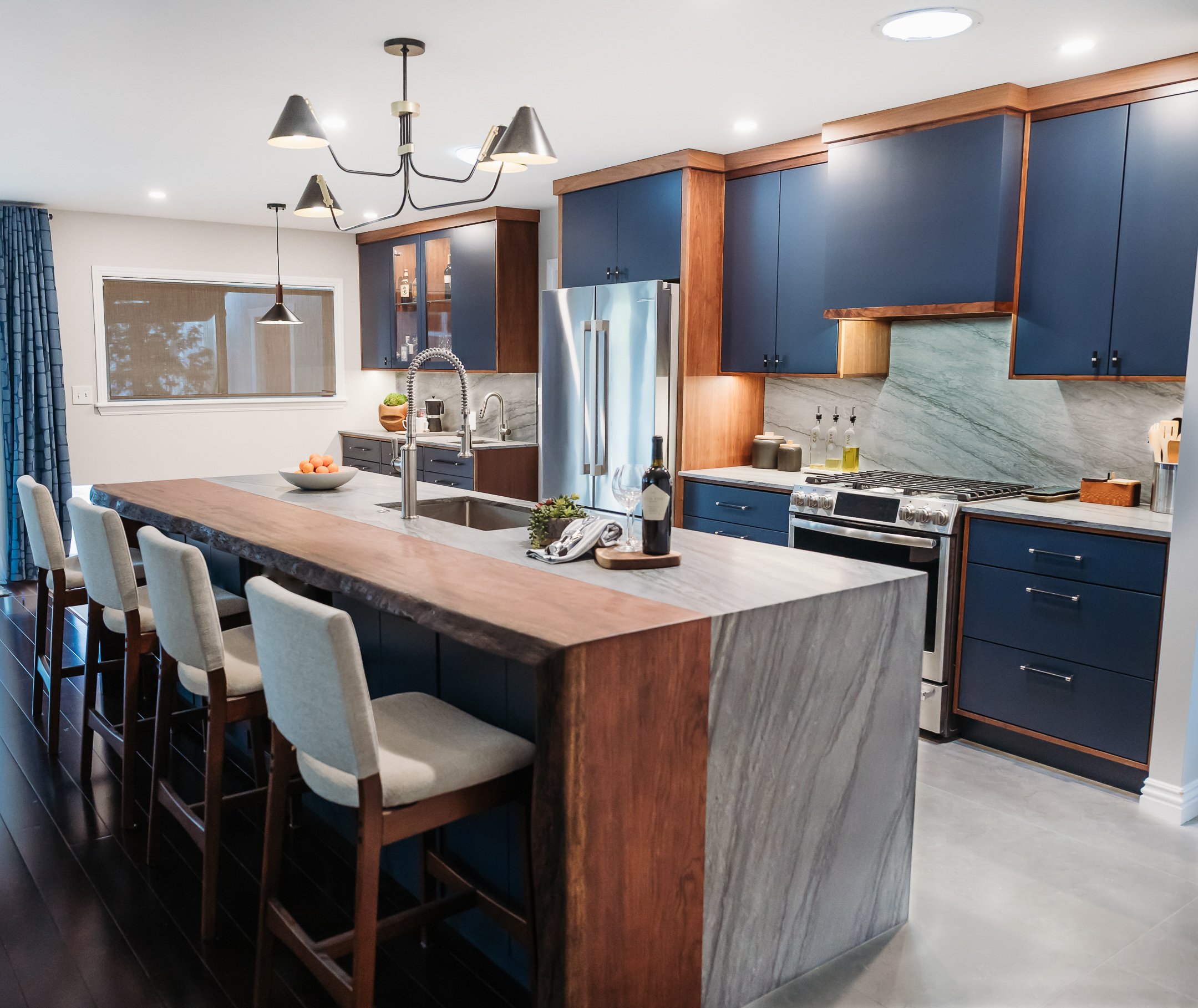Modern kitchen with blue cabinets, marble backsplash, and a large wooden island with seating. Contains stainless steel appliances, a sink with a gooseneck faucet, and decorative items like bottles and a bowl of fruit.