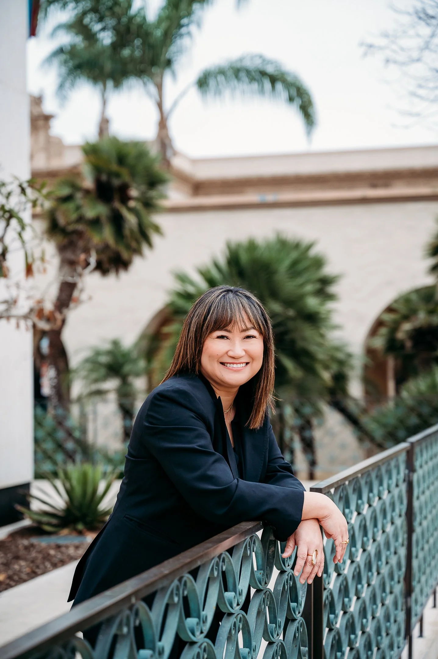 Salt Air Design Studio Principal Design Cain Luong stands leaning over a railing smiling wearing a black suit in Balboa Park