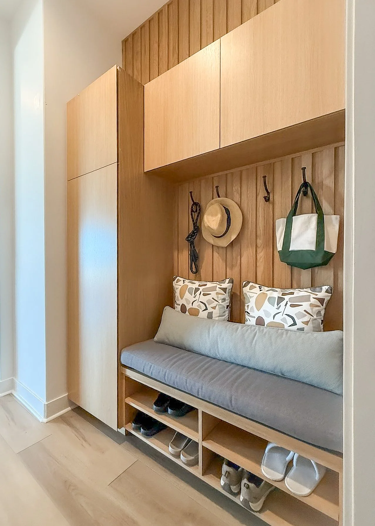 white oak built in mud room shoe storage cleaning closet upholstered bench black metal hooks denim cushion geometric pattern playful pillows light green lumbar