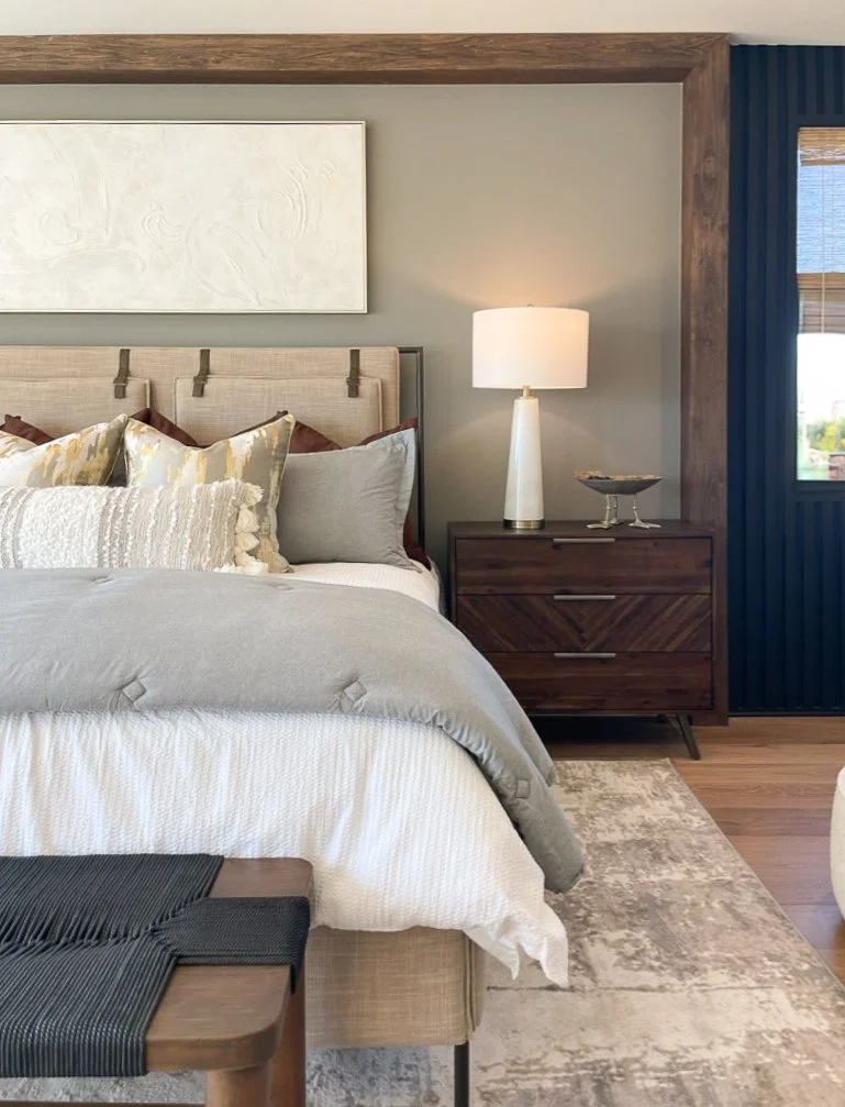 A bedroom with a beige upholstered bed, decorative pillows, and a gray and white bedding set, next to a wooden nightstand with a white table lamp and a glass dish, situated in a room with a textured wall and a window with blinds.