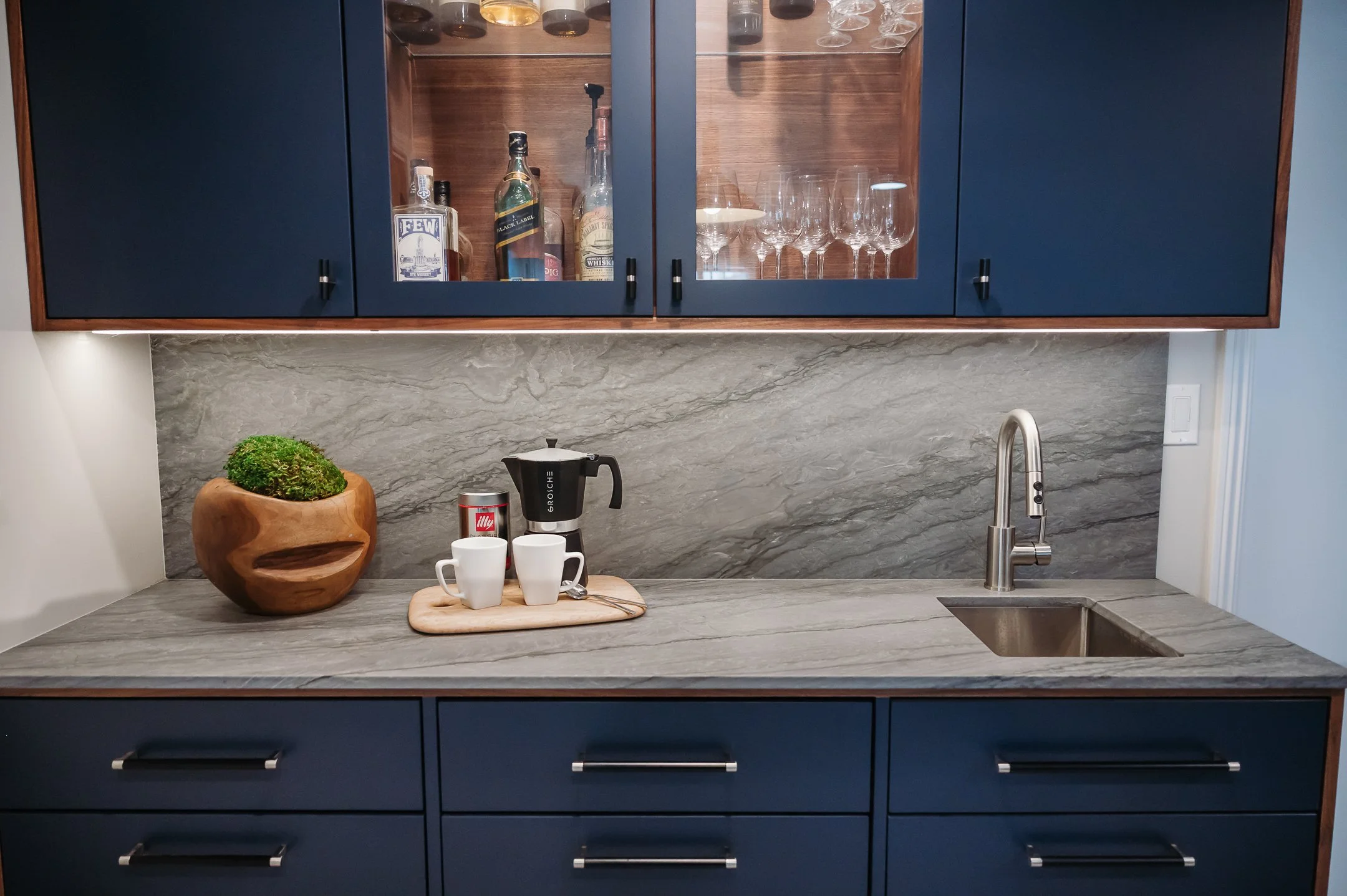 A kitchen with dark blue cabinets, a gray marble countertop, and a matching gray marble backsplash. There is a stainless steel sink with a curved faucet on the right. On the countertop, there is a wooden tray holding two white coffee mugs, a spoon, a