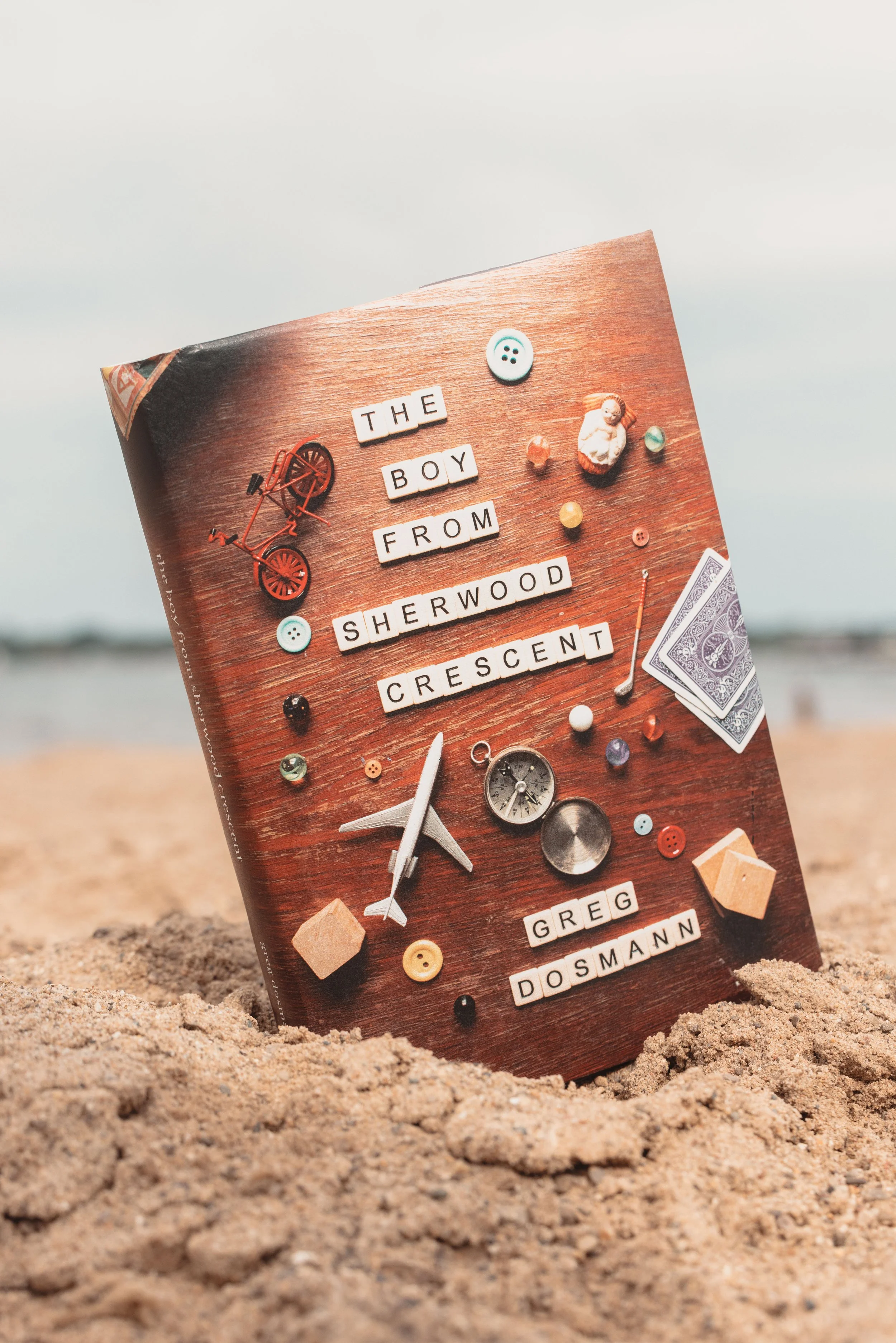 The book on a sandy beach. The book is partly buried in the sand with a cloudy sky in the background. The visible words on the cover spell out "The Boy from Sherwood Crescent by Greg Dosmann".